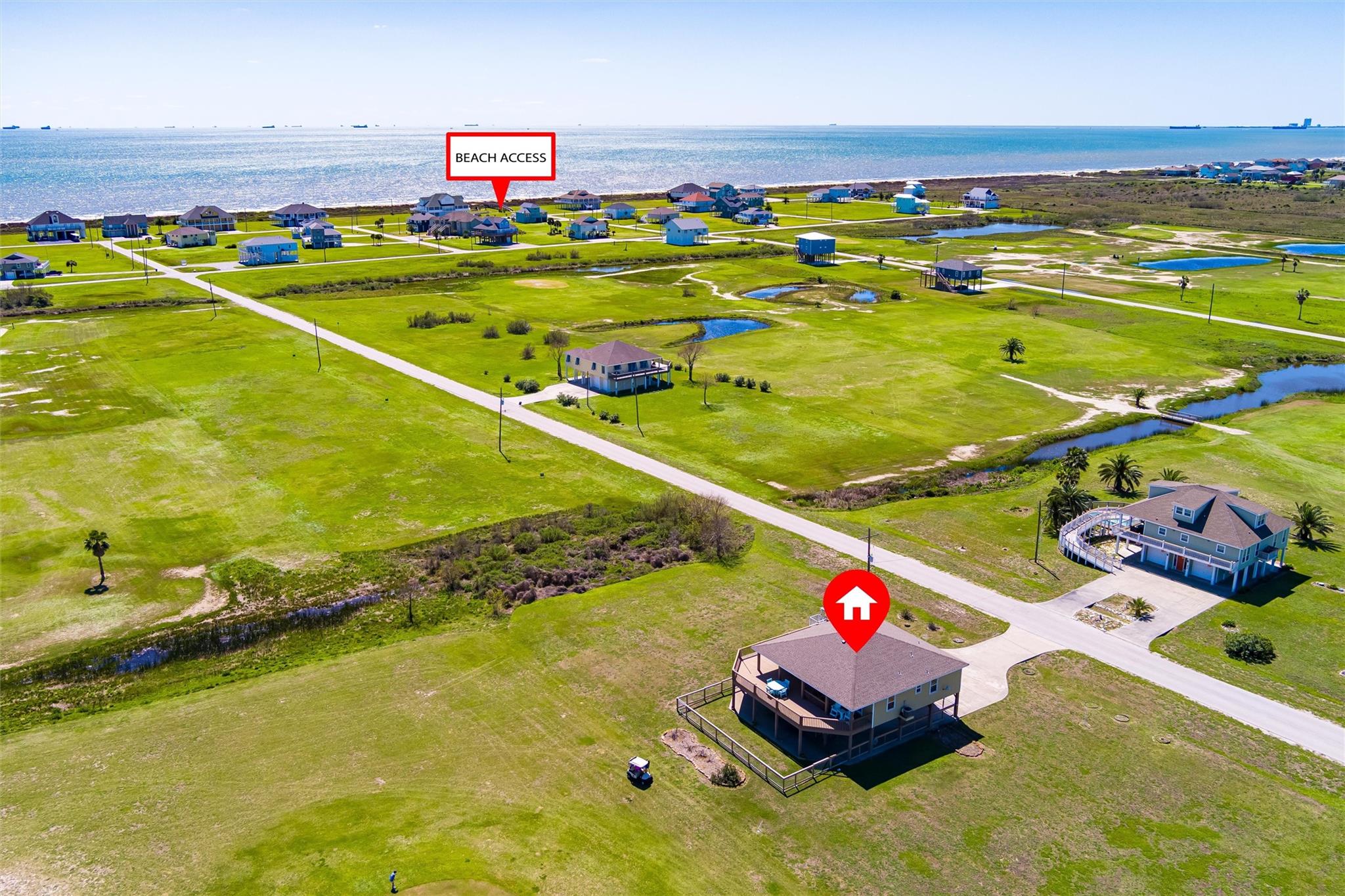 Aerial view of a raised coastal home near beach access, with open grassy lots and shoreline.