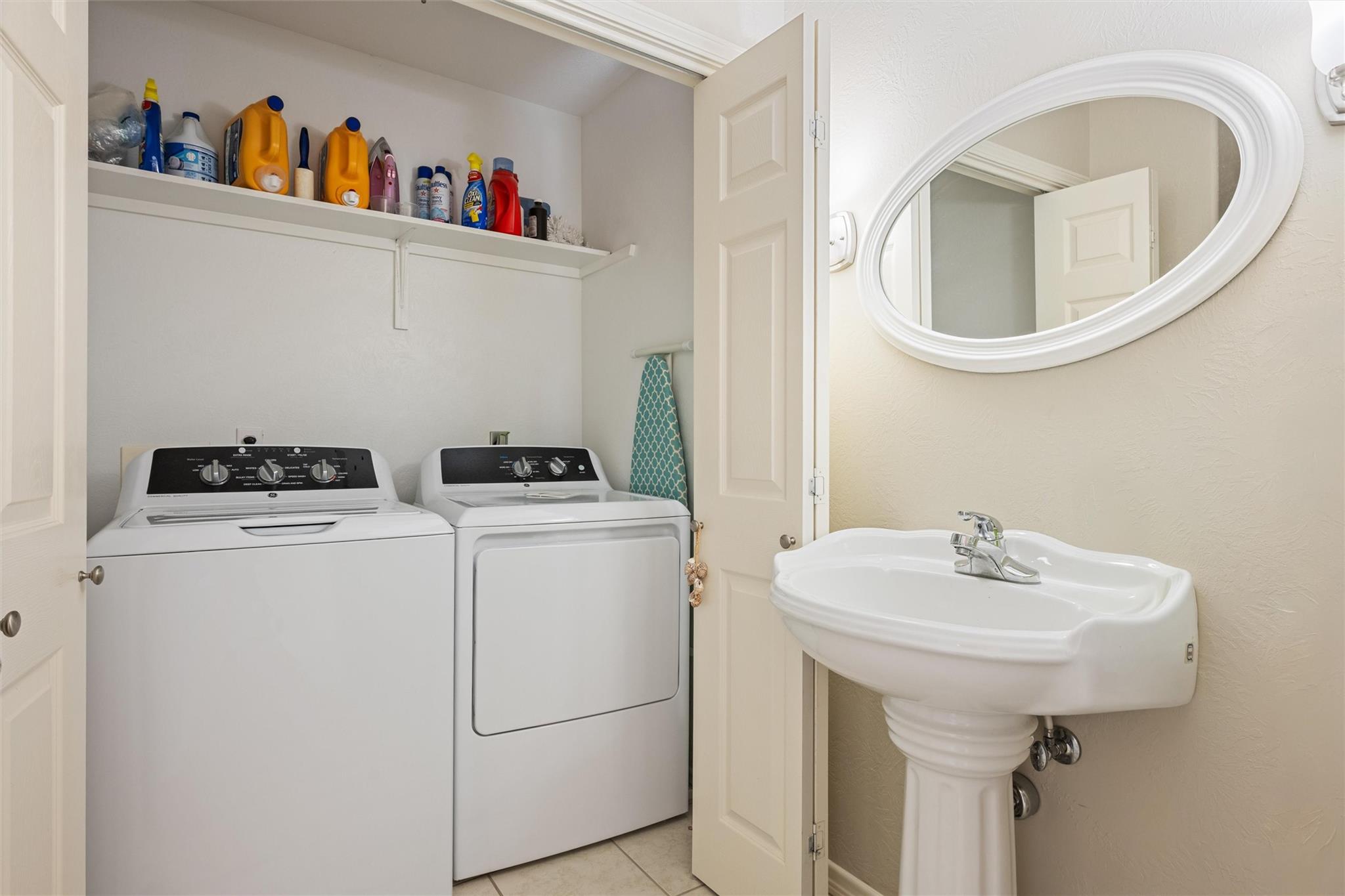 Laundry room with washer and dryer, cleaning-supply shelf, and pedestal sink beside an oval mirror.