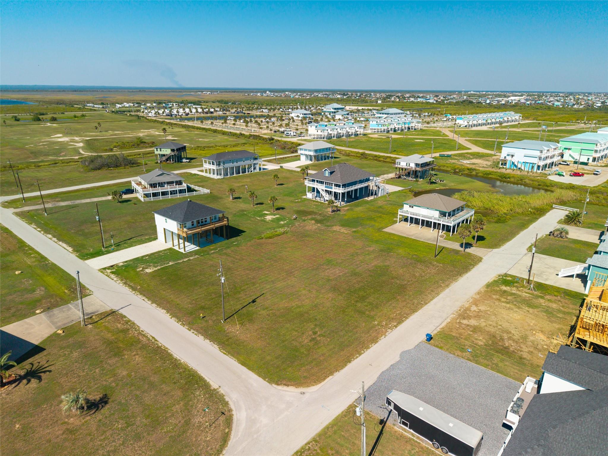Aerial view of open grassy lots among elevated coastal homes, with marshland and water in the distance.