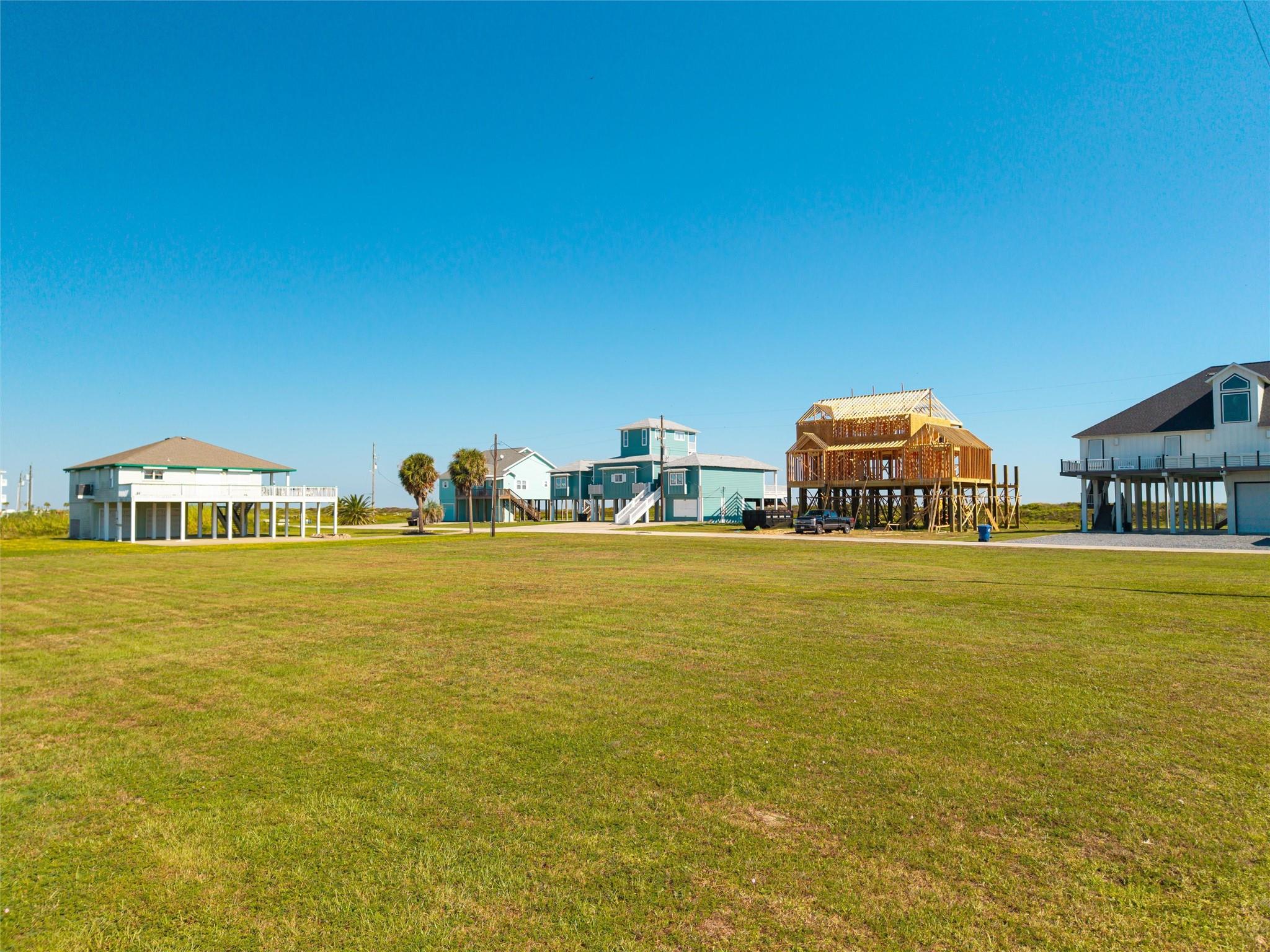 Elevated coastal homes beside a broad grassy lot, including a two-story house under construction.