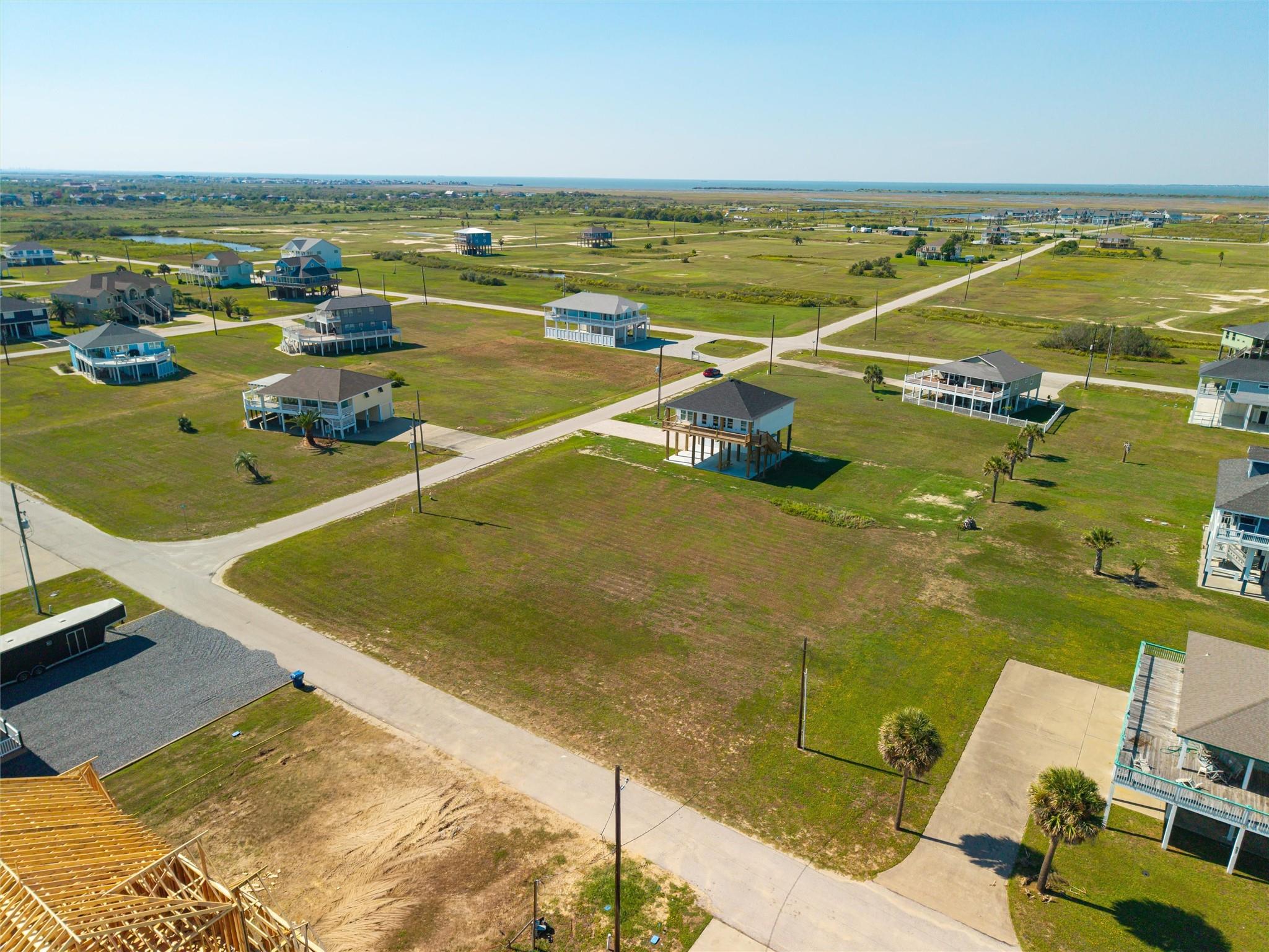 Aerial view of a coastal neighborhood with elevated homes, open lots, and distant water.