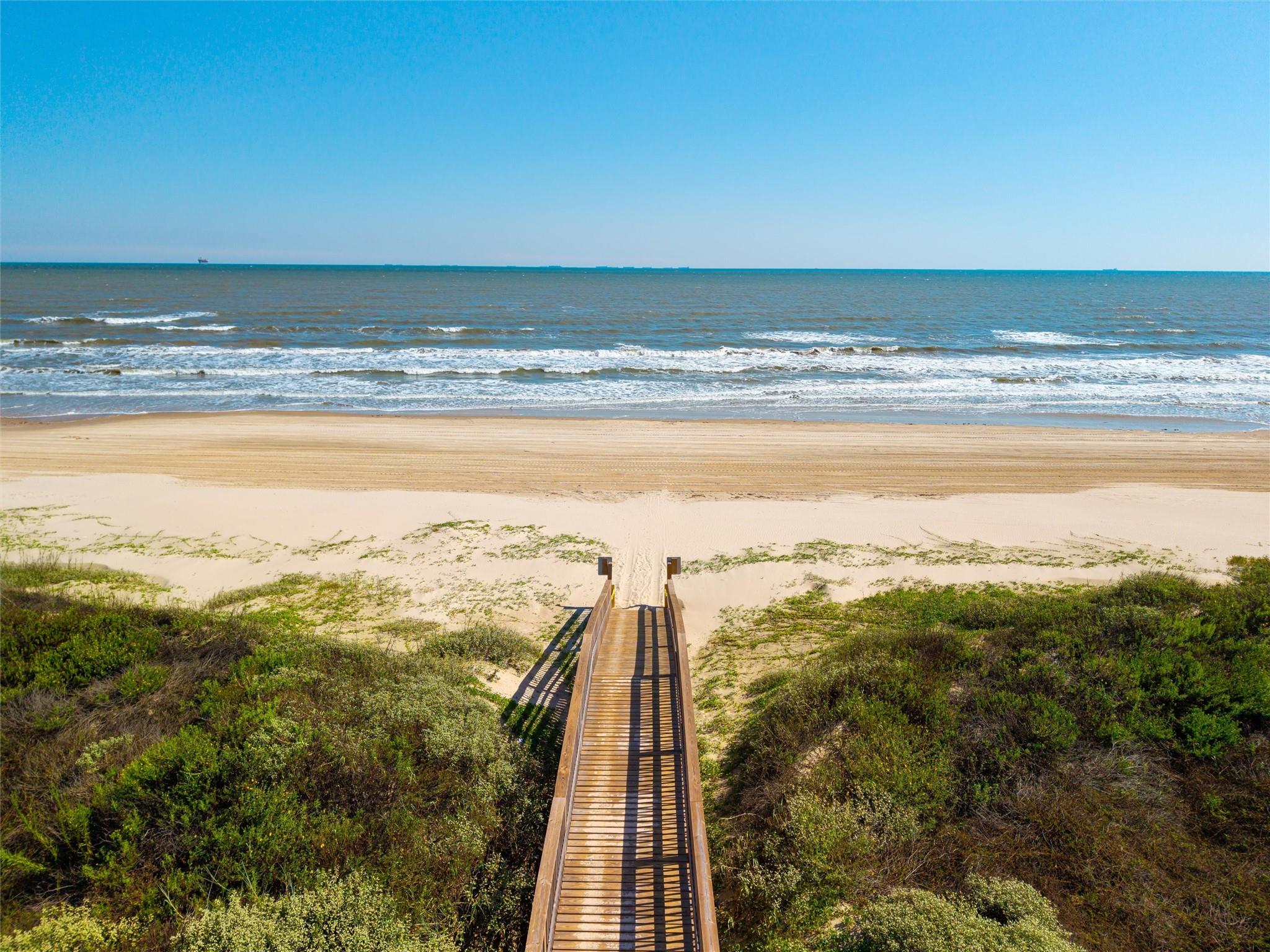A wooden boardwalk crosses coastal vegetation to a sandy beach and ocean.
