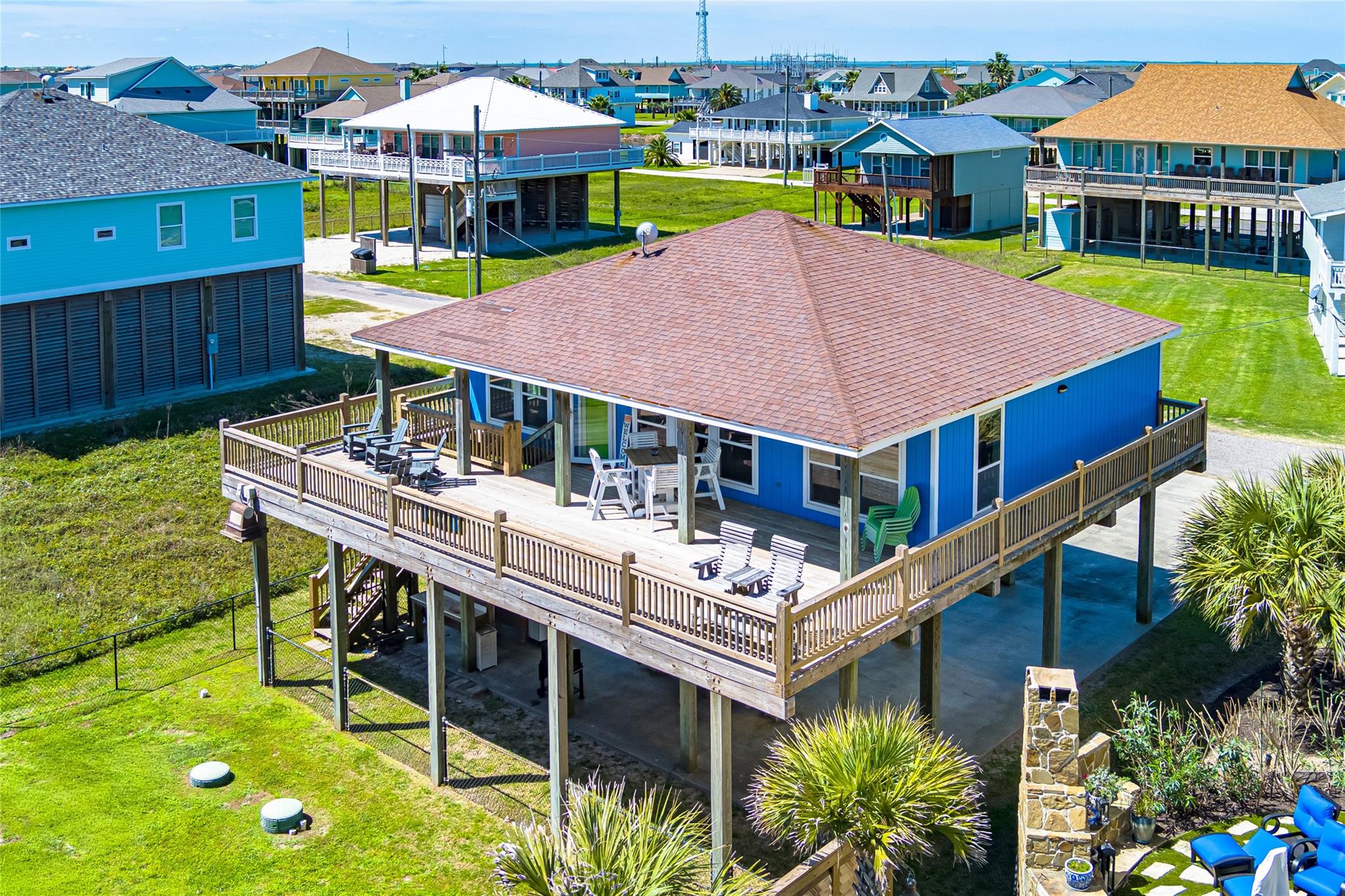 Elevated blue coastal house with a wide furnished wraparound deck among neighboring beach homes.