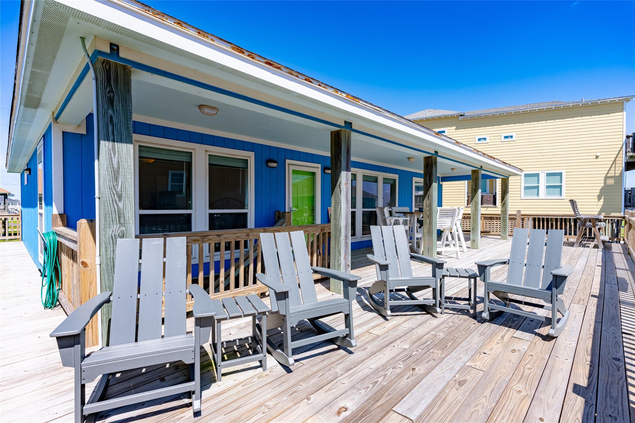 Blue coastal house with a covered wooden deck furnished with gray Adirondack chairs.
