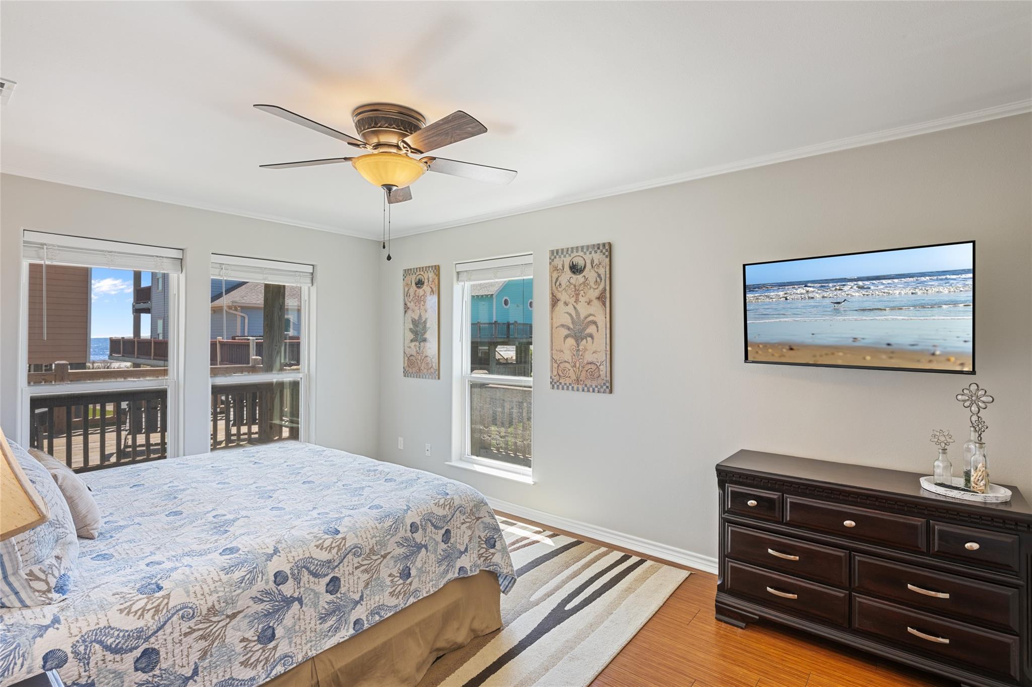 Bedroom with a patterned bed, dark dresser, and windows overlooking neighboring homes and a glimpse of water.