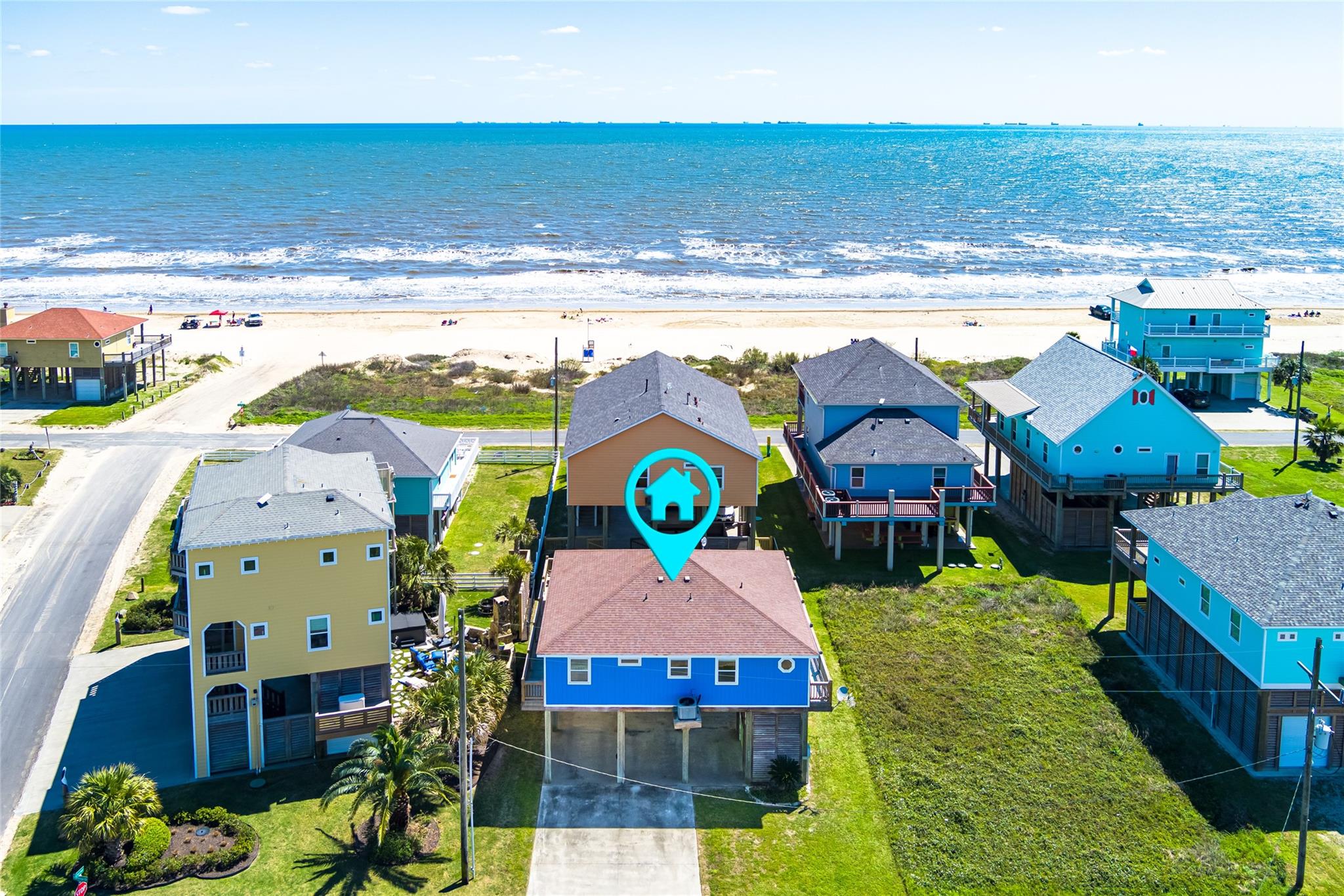 Aerial view of a blue elevated beach house near the shoreline, marked with a turquoise home pin.