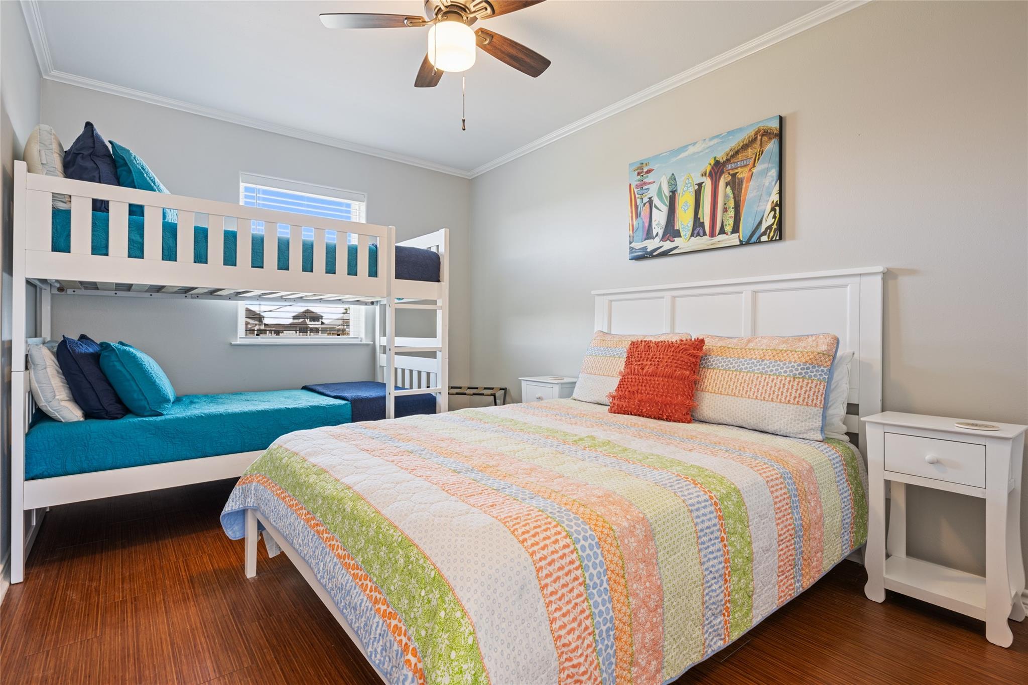 Bedroom with a white bunk bed, colorful quilts, surfboard artwork, and dark wood flooring.