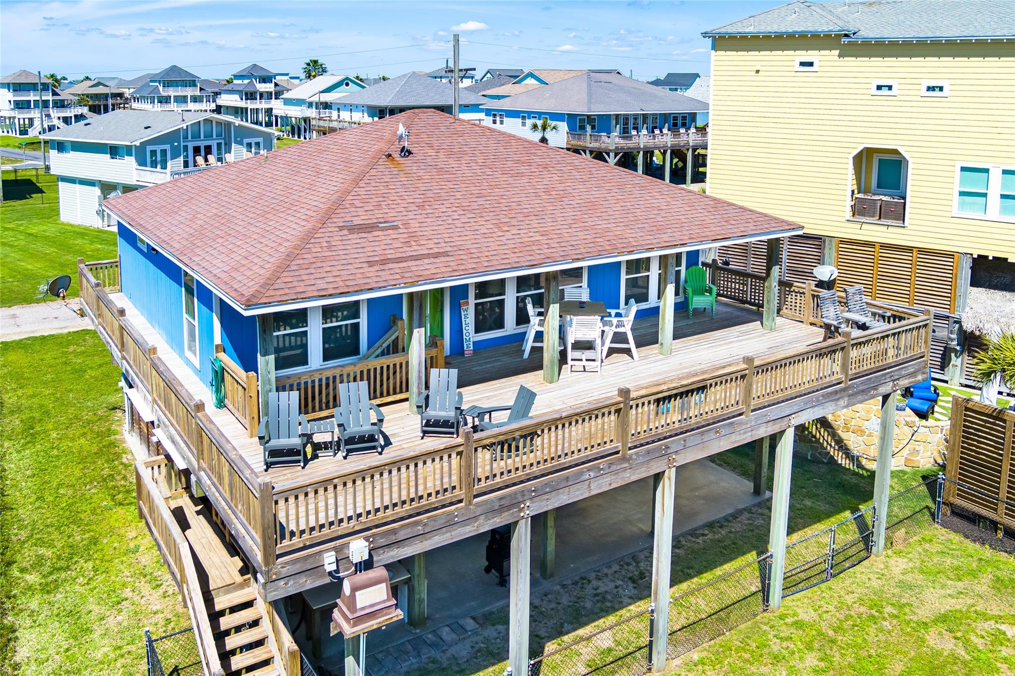 Blue elevated coastal home with a large furnished wooden deck and neighboring beach houses.