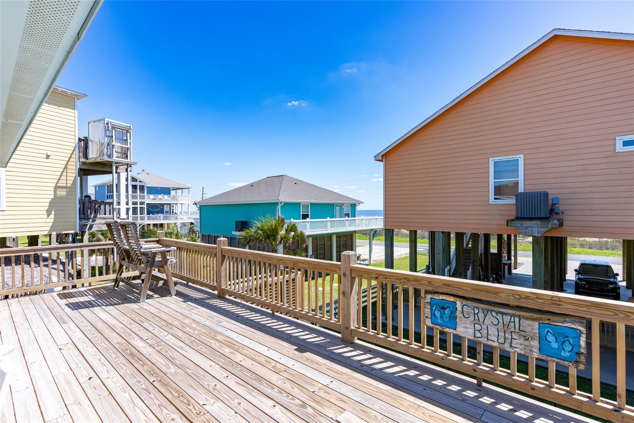 Wooden deck with outdoor chairs and a Crystal Blue sign, overlooking elevated coastal homes and a glimpse of water.