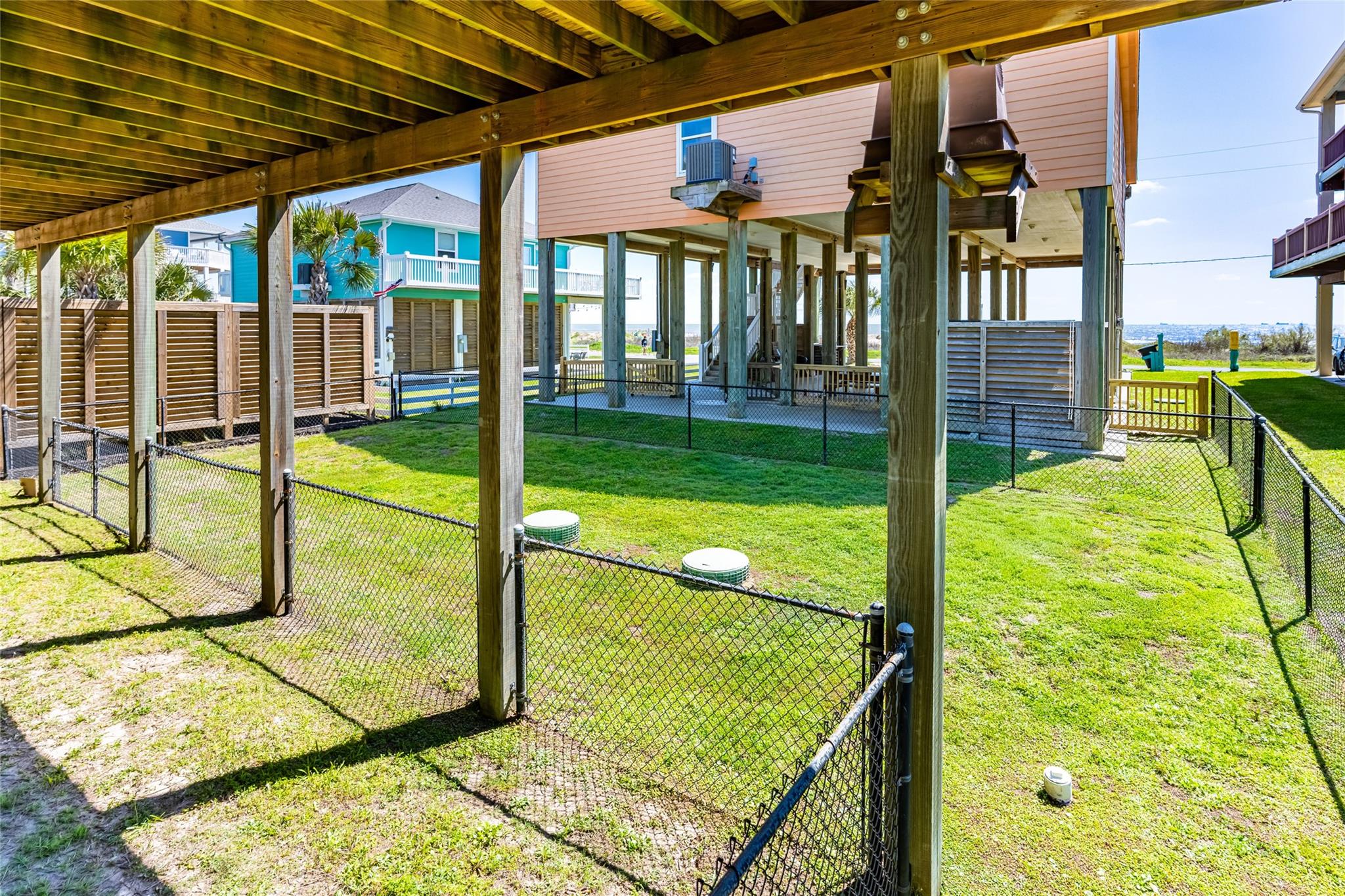 Fenced grassy backyard beneath an elevated beach house, with neighboring homes and ocean visible beyond.