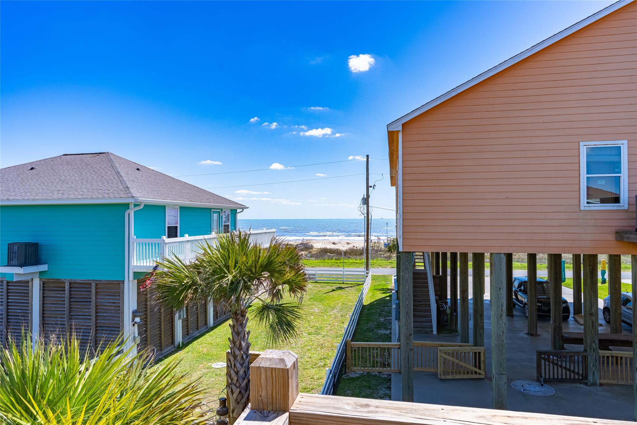 Ocean view between raised beach houses, with a palm tree and grassy yards.
