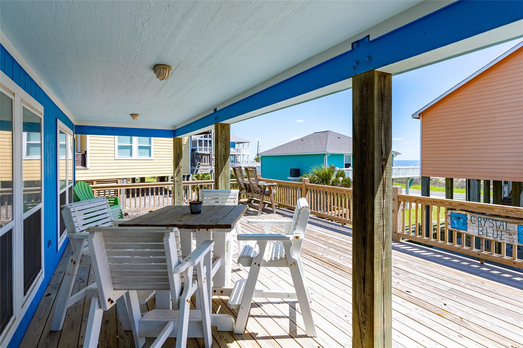 Covered beach-house deck with white Adirondack chairs, a dining table, and a glimpse of the water.