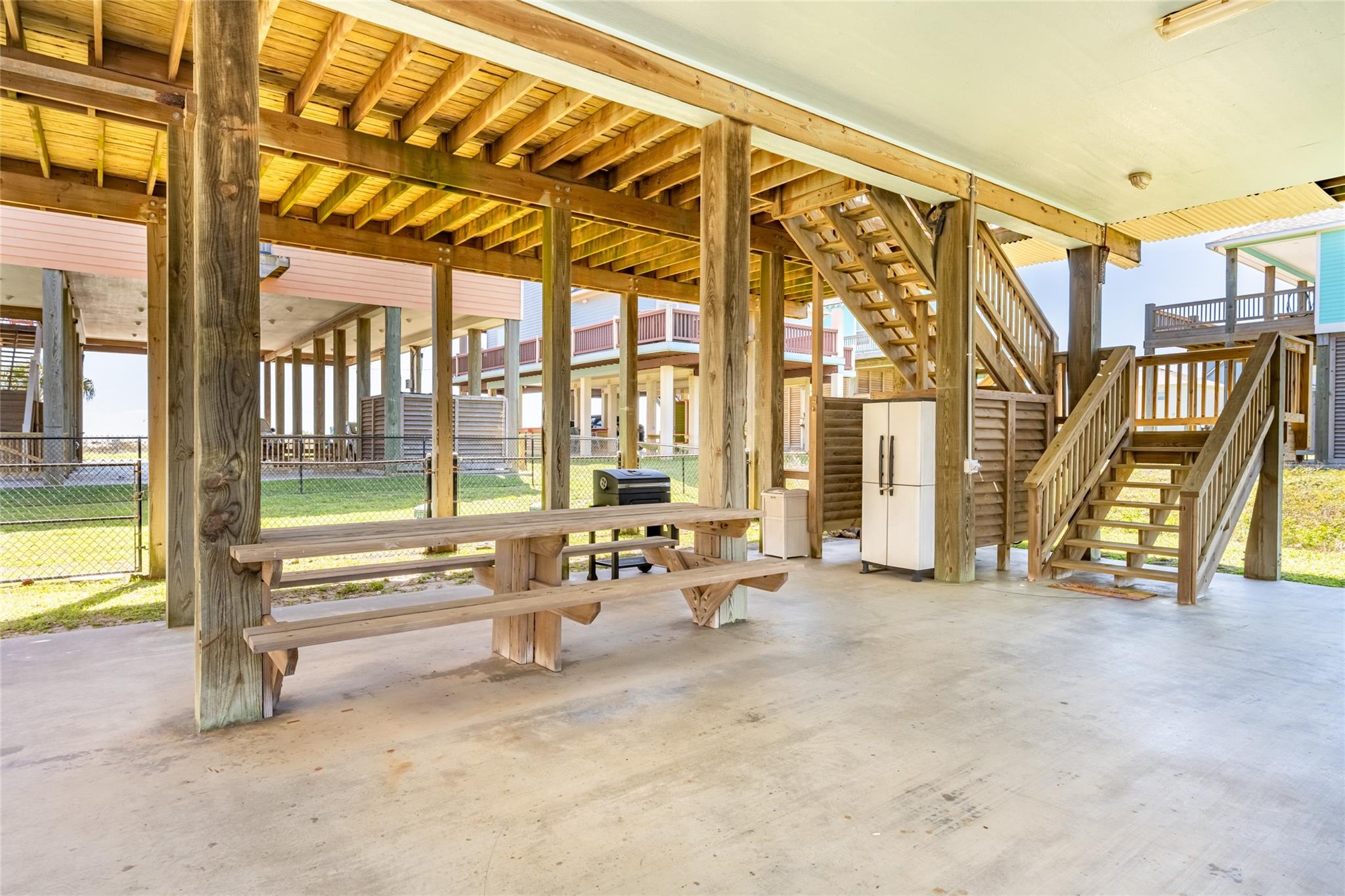 Covered patio beneath a raised home with a picnic table, refrigerator, grill, and wooden stairs.