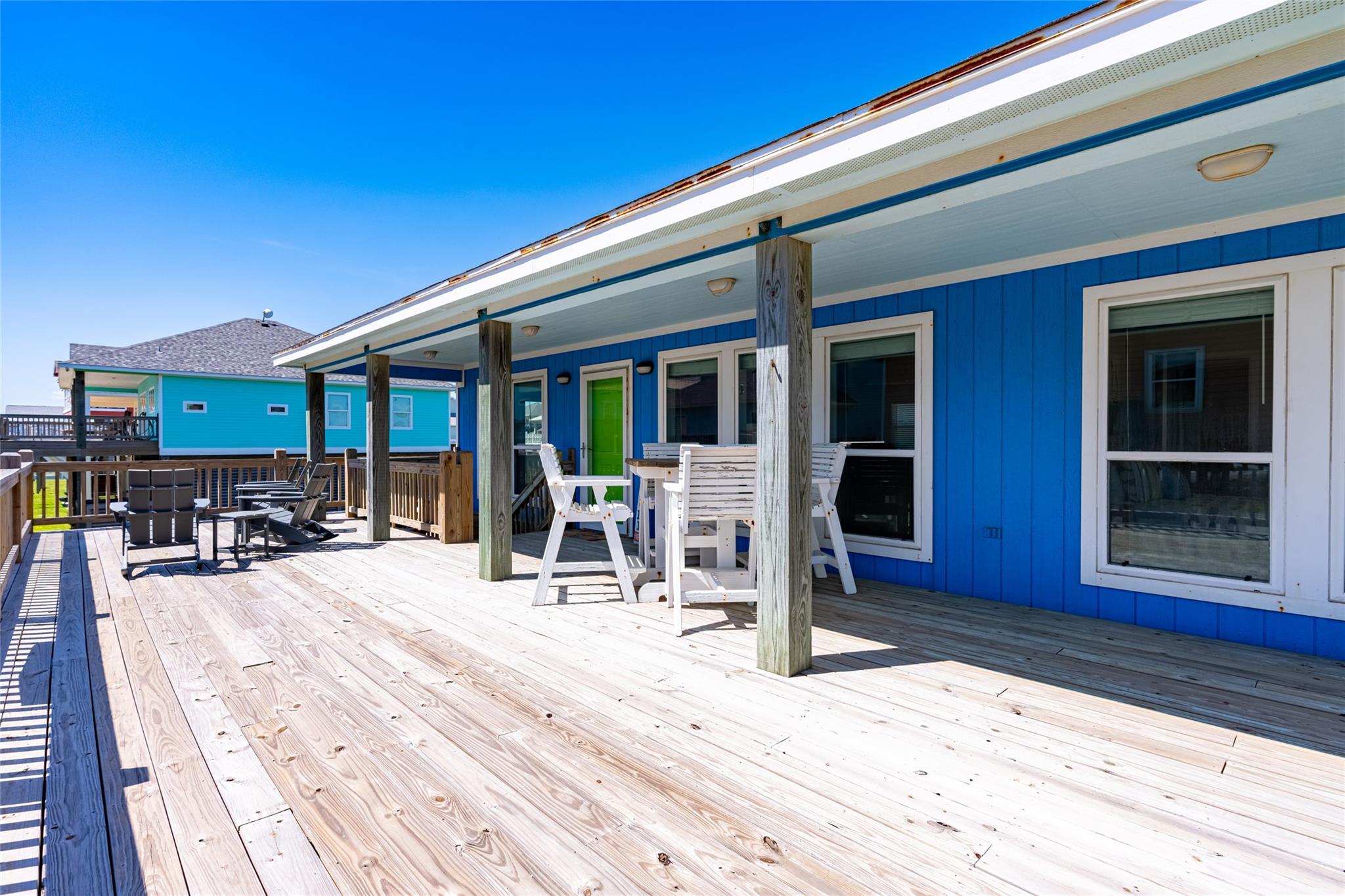 Wide wooden deck beside a bright blue house, with a covered porch and white outdoor dining furniture.