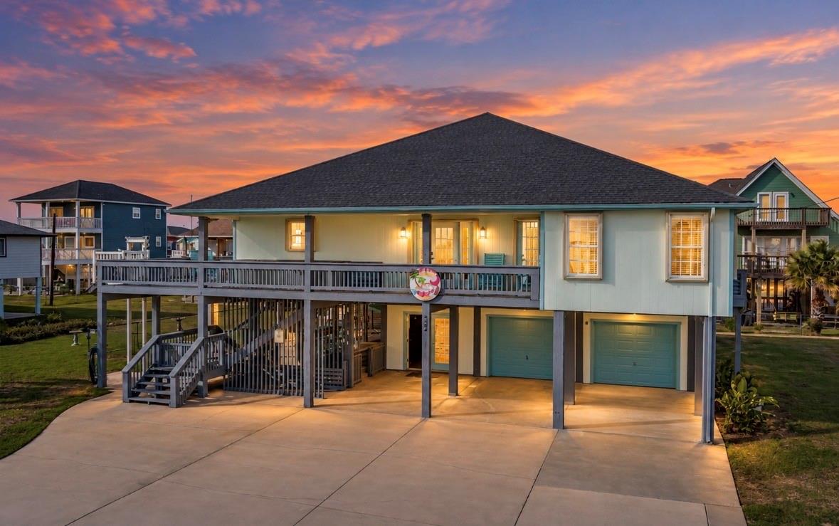 Raised coastal house with a wraparound deck and two garage doors under a pink-orange sunset.