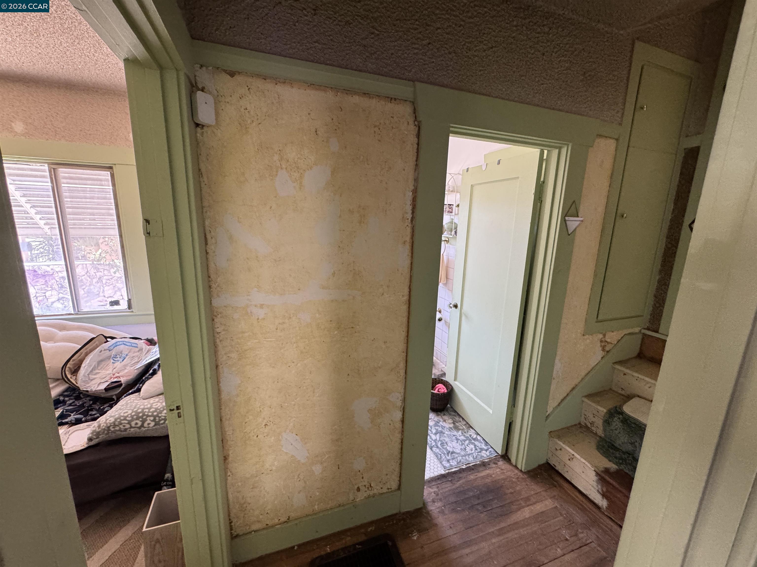 Hallway with worn patched walls, stairs, and open doorways to a bedroom and bathroom.