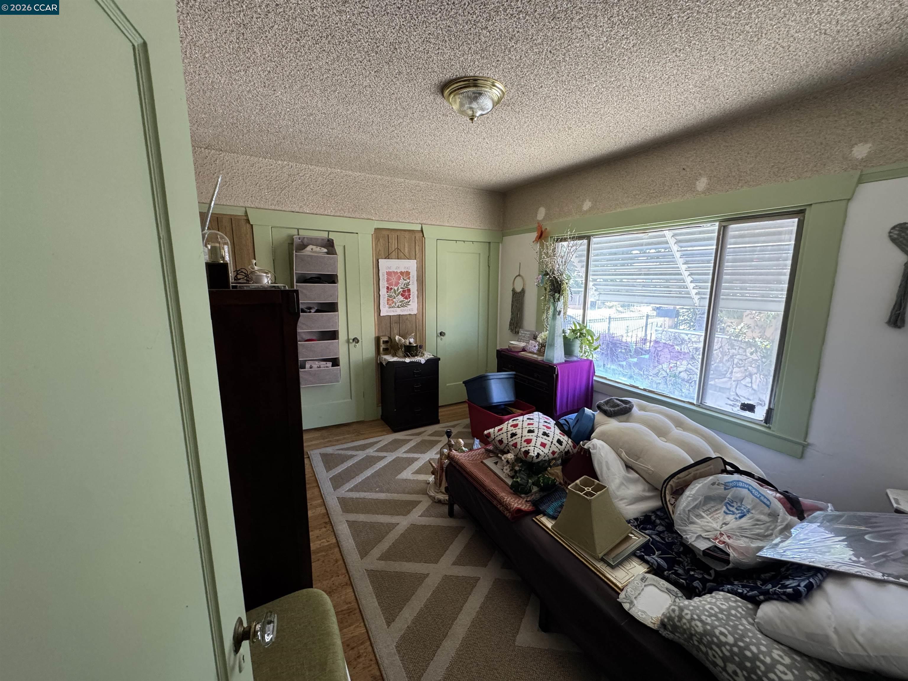 Bedroom with pale green paneled walls, a large shuttered window, and a daybed among assorted belongings.