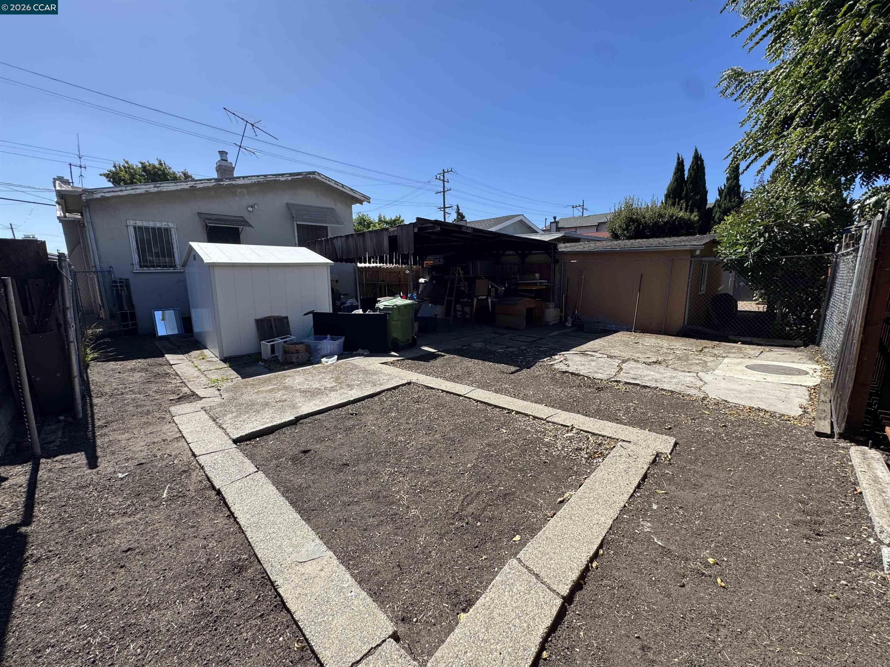 Fenced backyard with a white shed, covered storage area, and concrete edging around bare soil.
