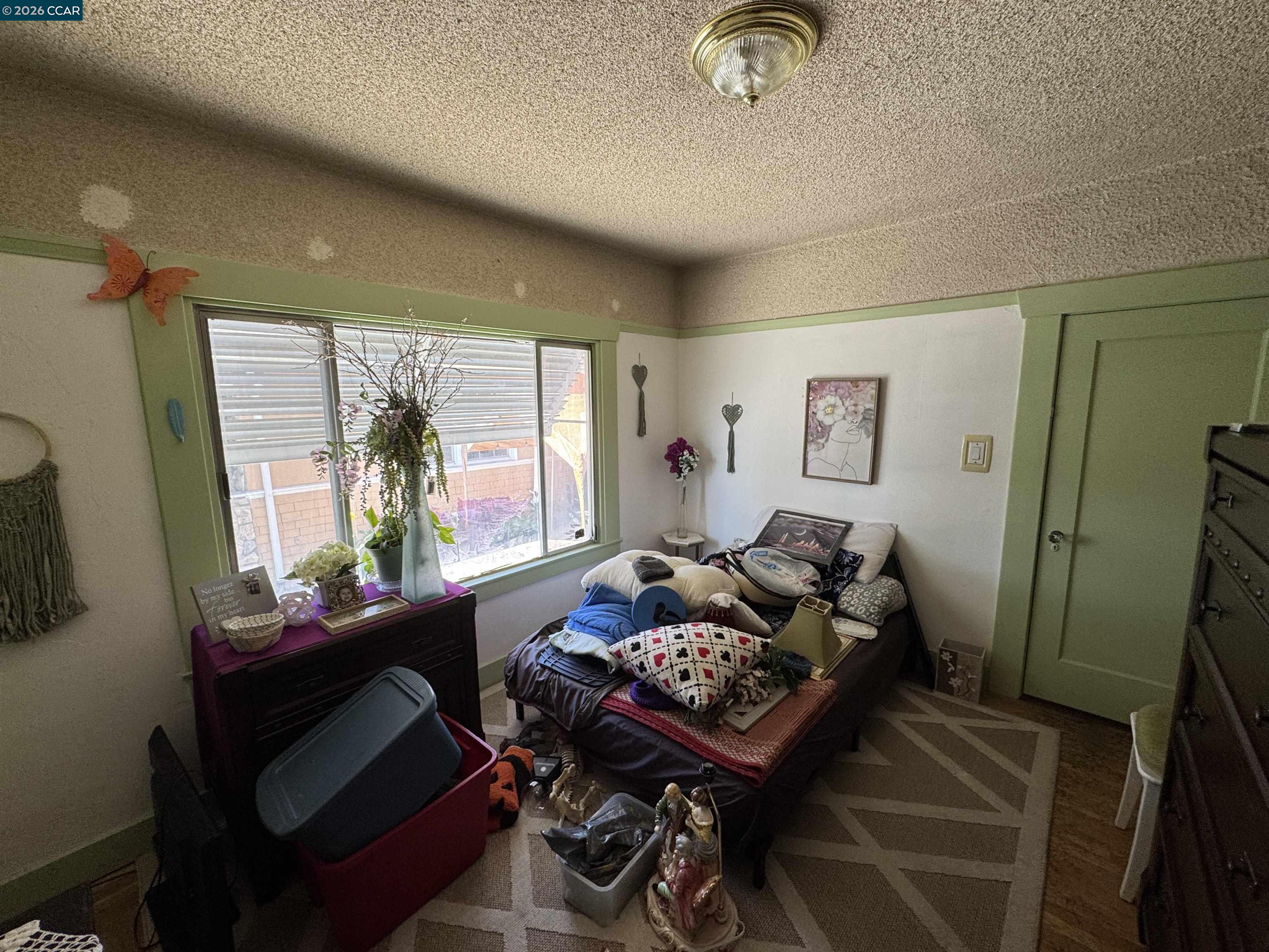 Crowded bedroom with green trim, a large window, and a bed piled with pillows and belongings.