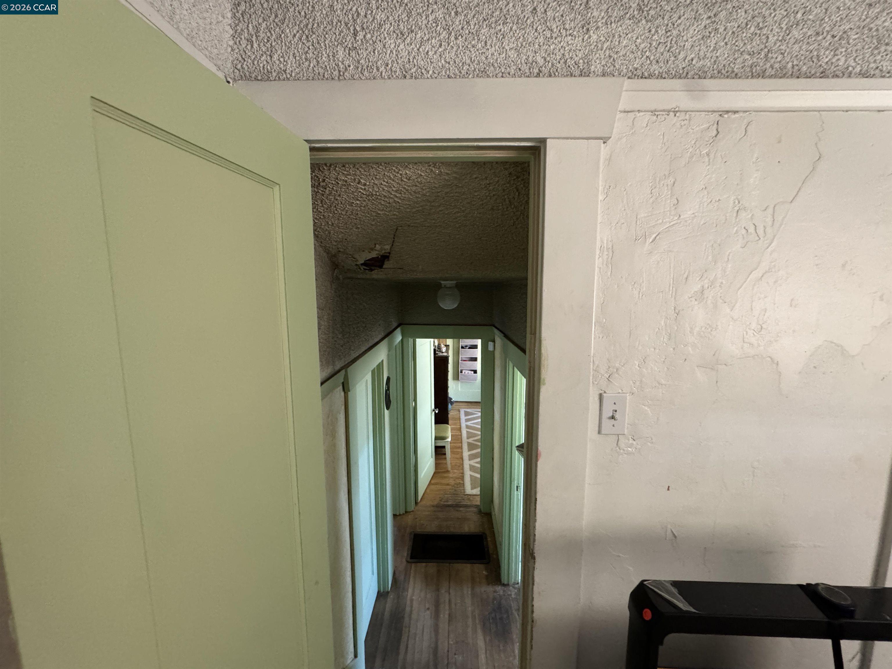 Narrow interior hallway with green-trimmed doorways, wood flooring, and damaged ceiling.