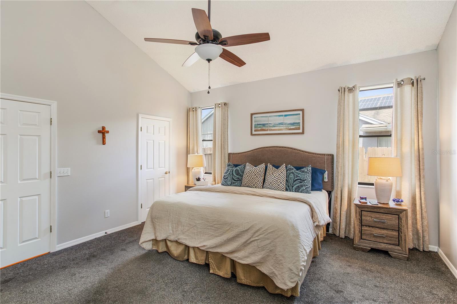 Furnished bedroom with a vaulted ceiling, ceiling fan, and bed between curtained windows.