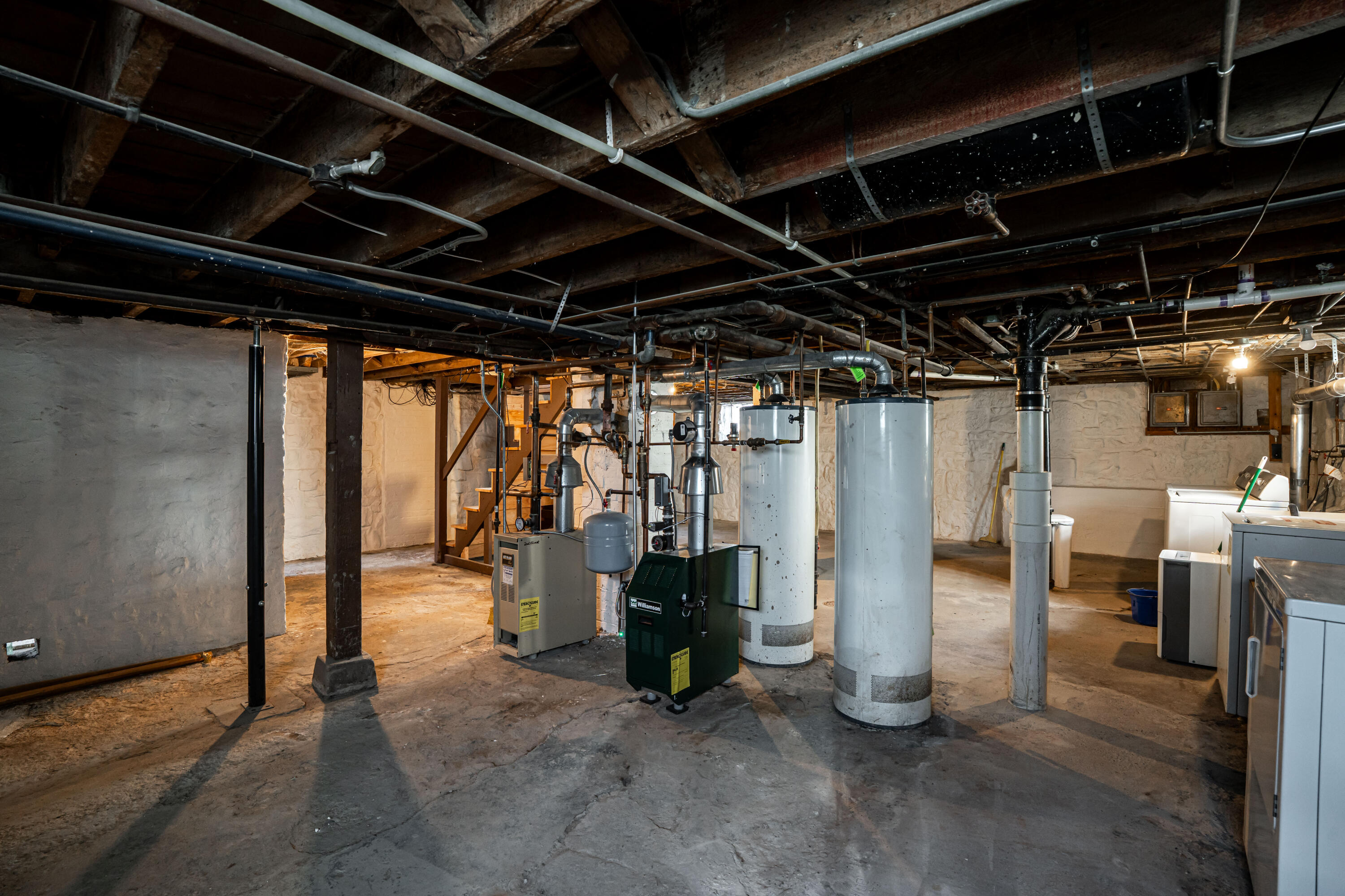 Basement mechanical room with boilers, water heaters, exposed pipes, and laundry machines.