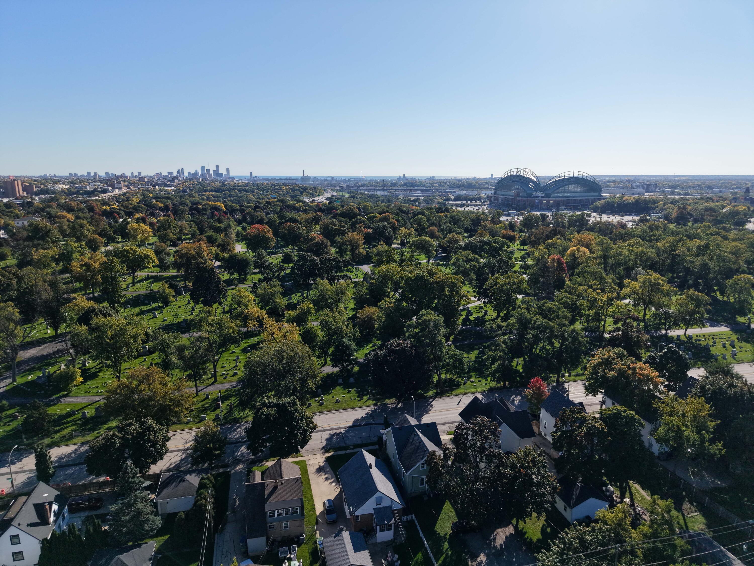 Aerial view of homes beside a tree-filled cemetery, with the Milwaukee skyline and American Family Field beyond.