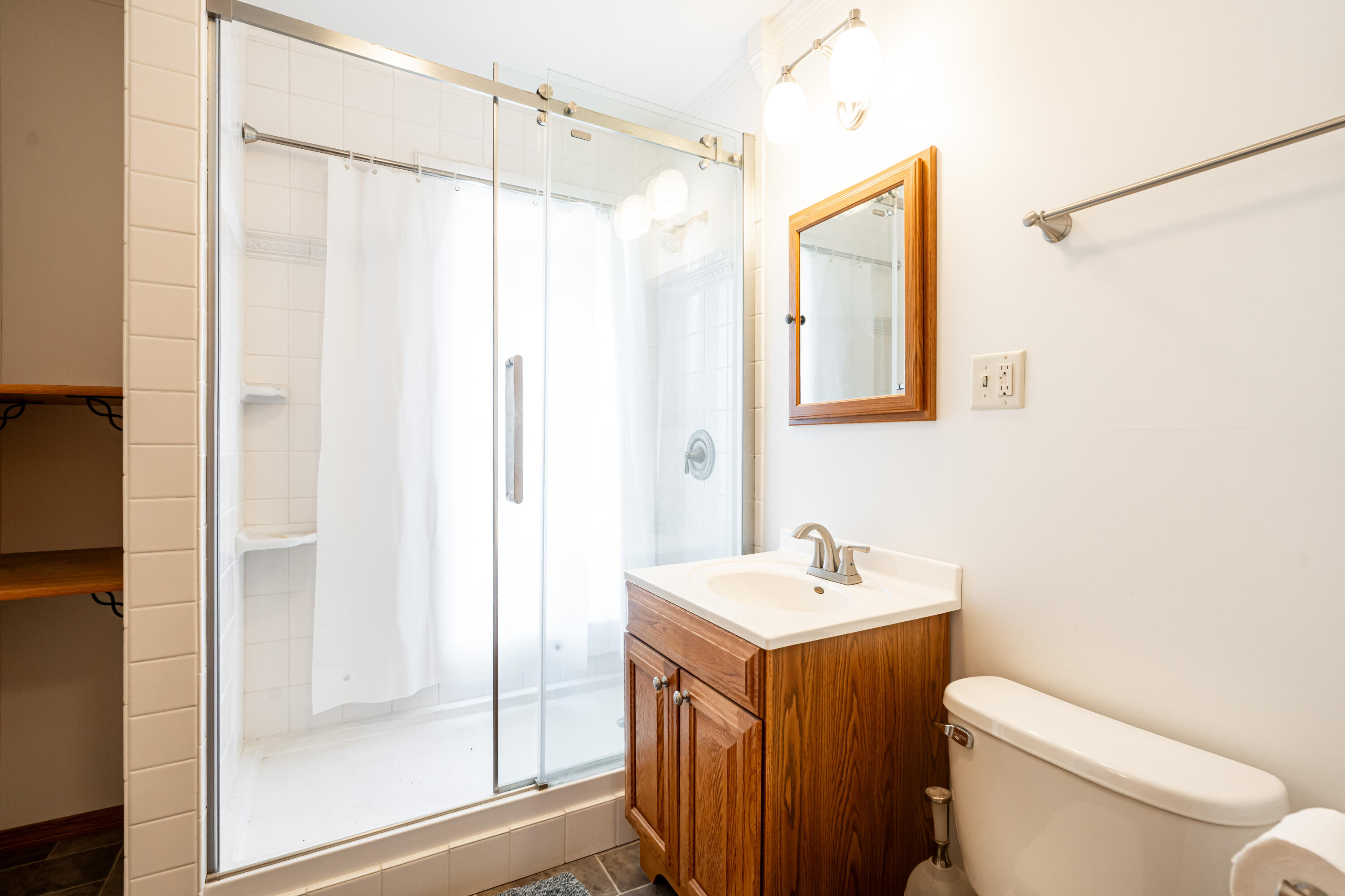 Bathroom with glass shower, white tile, and a wood vanity beside the toilet.