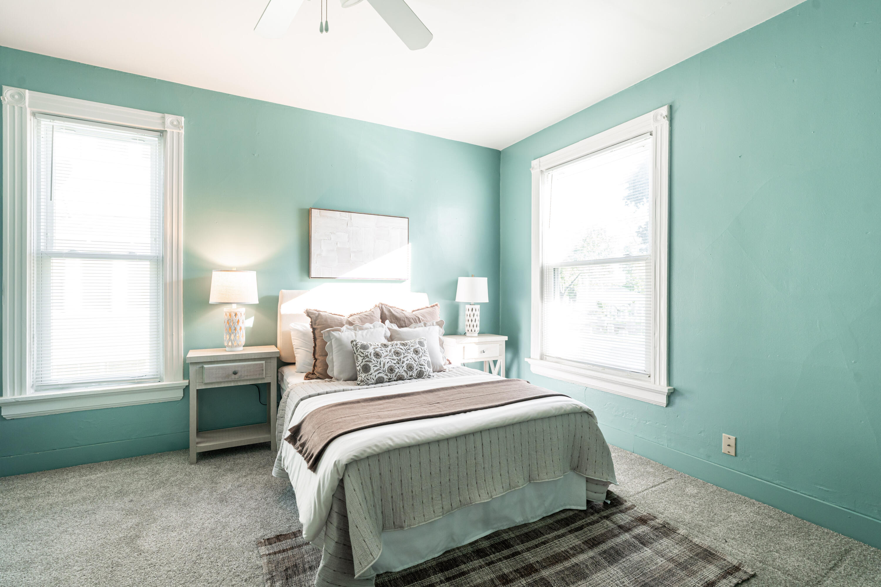 Staged bedroom with aqua walls, a neatly made bed, and two bright windows.