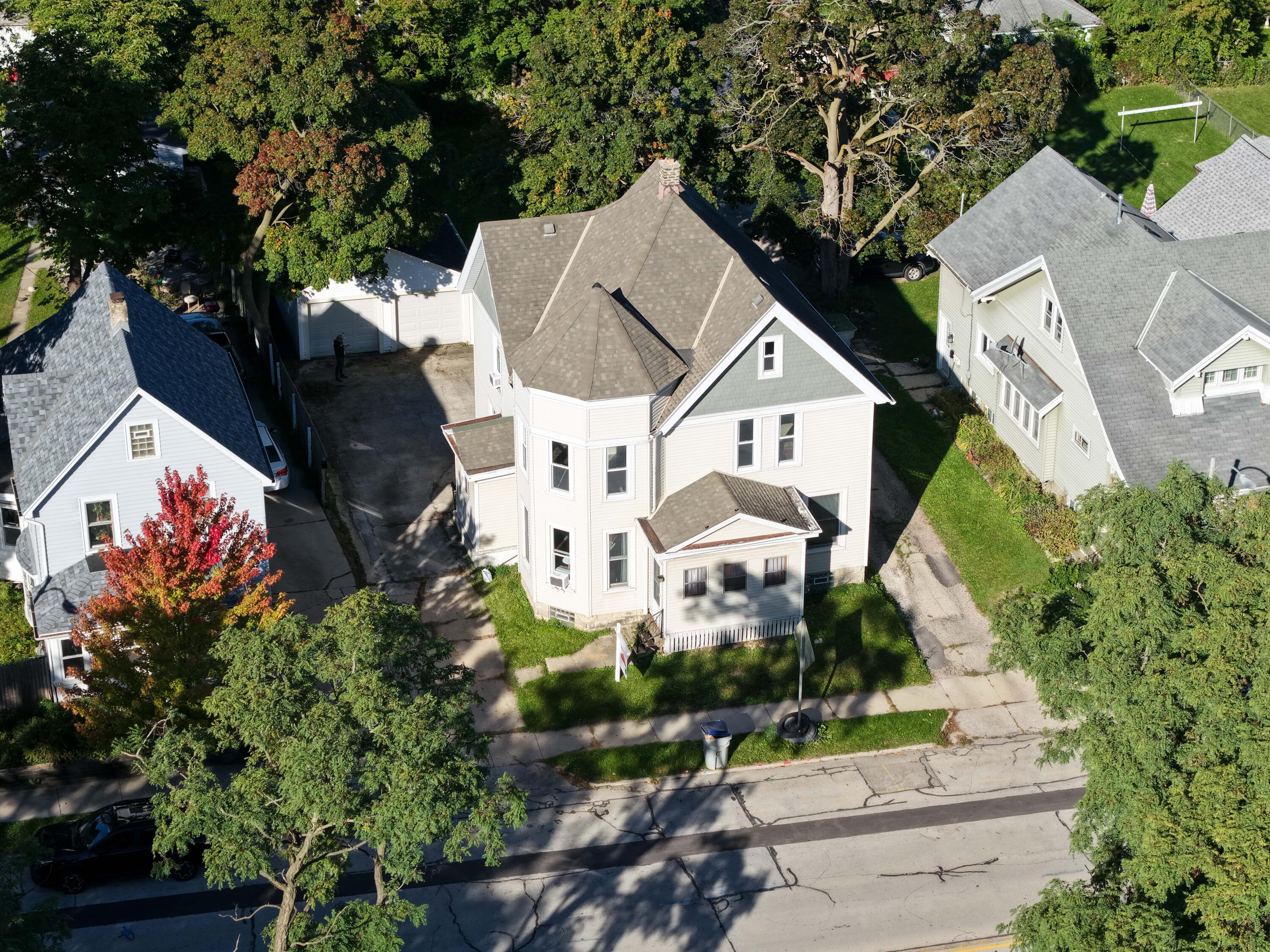 Aerial view of a light-colored two-story house with a steep gray roof and bay windows, surrounded by mature trees.