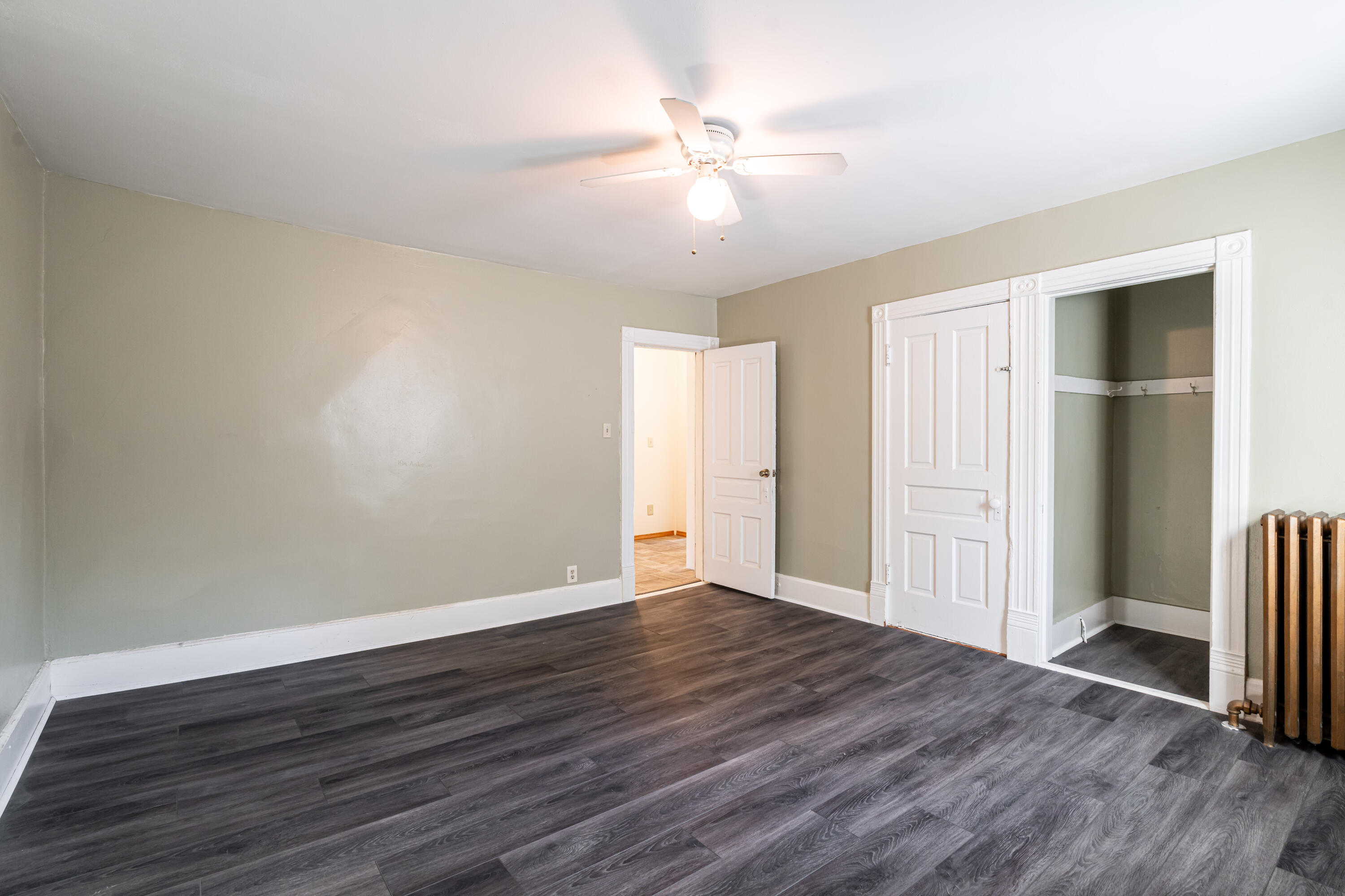 Empty room with dark plank flooring, a ceiling fan, decorative white trim, and a cast-iron radiator.