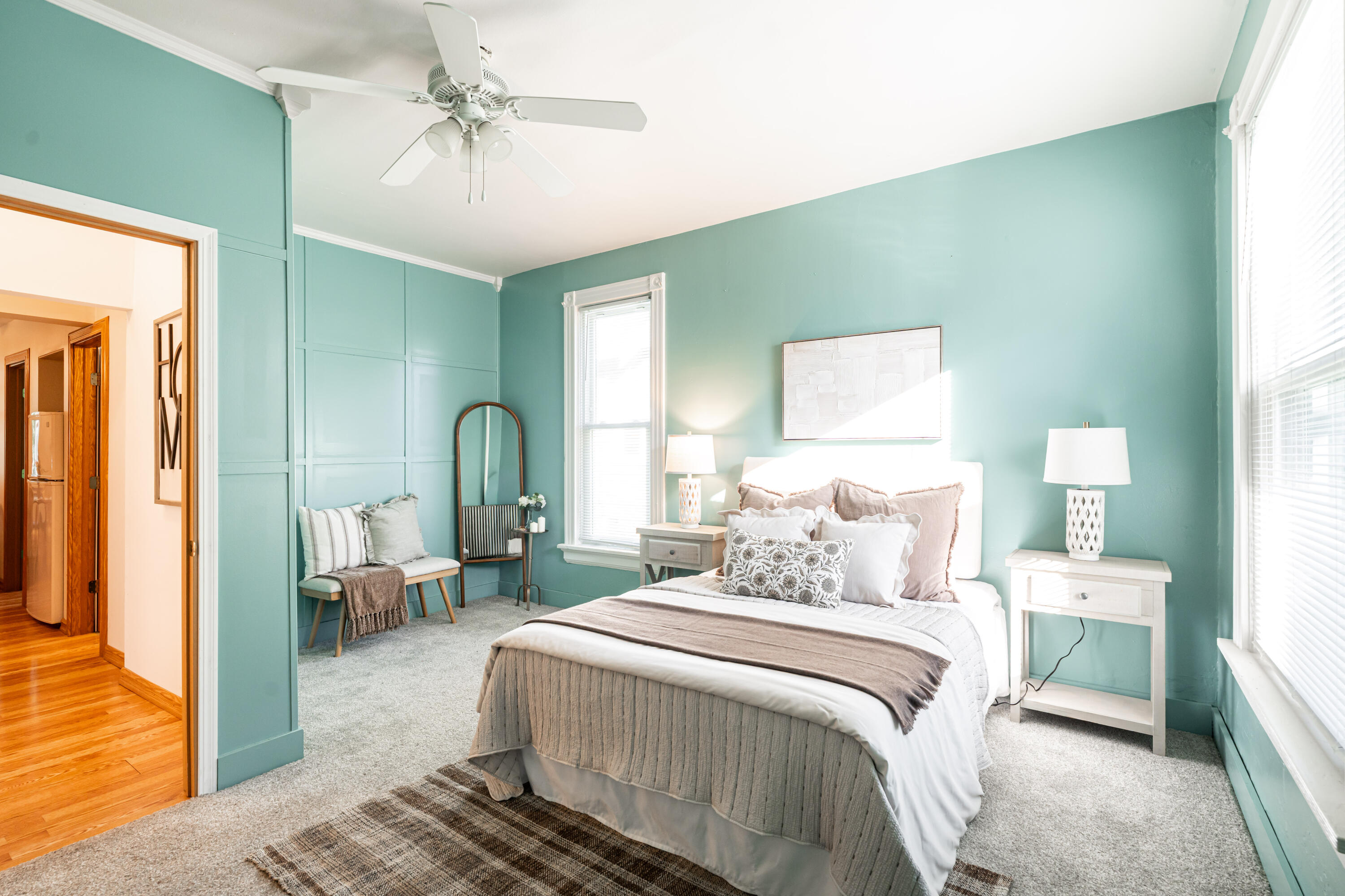 Staged bedroom with teal paneled walls, a neutral bed, an arched mirror, and a ceiling fan.