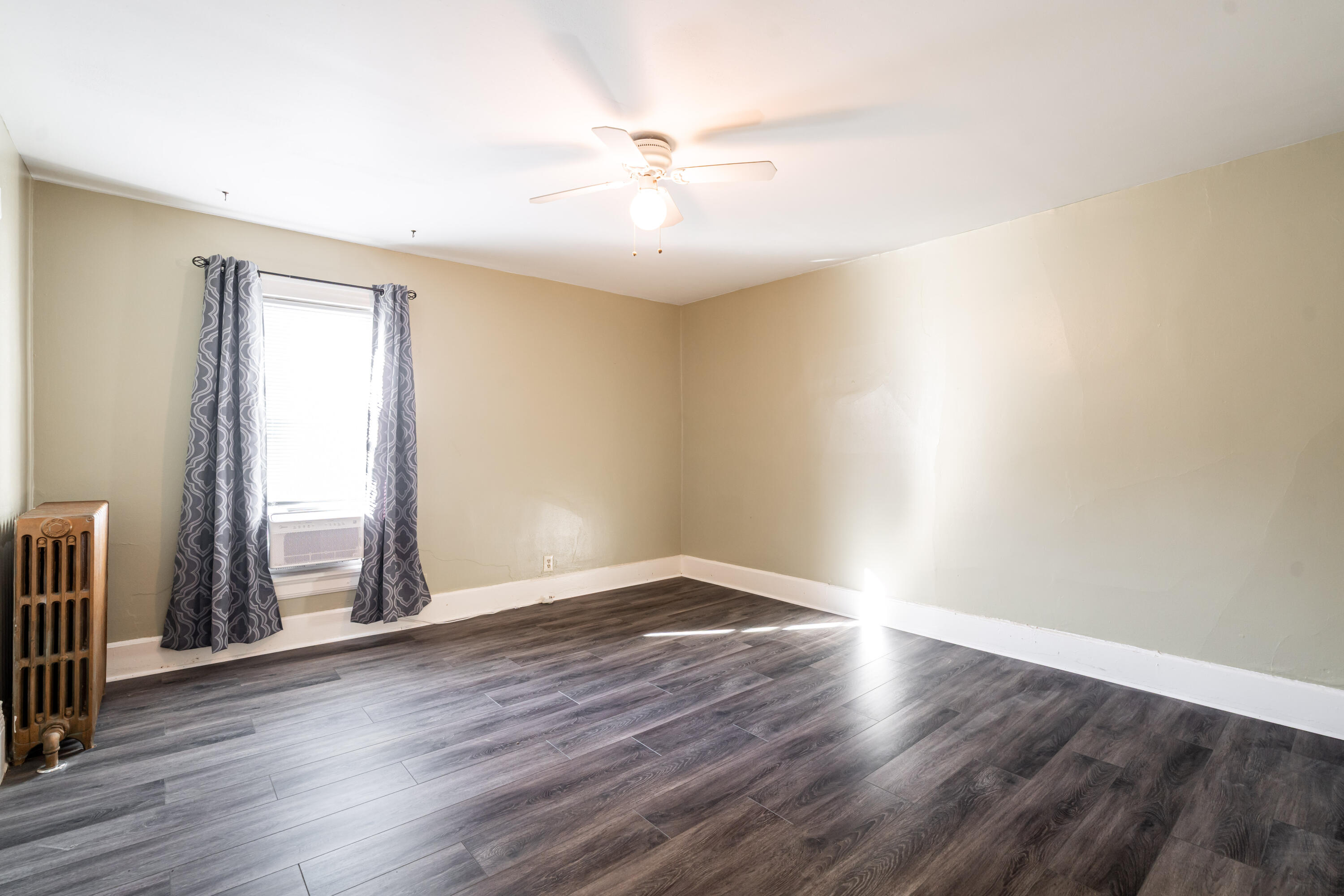 Empty room with dark wood-look floors, patterned curtains, window air conditioner, and radiator.