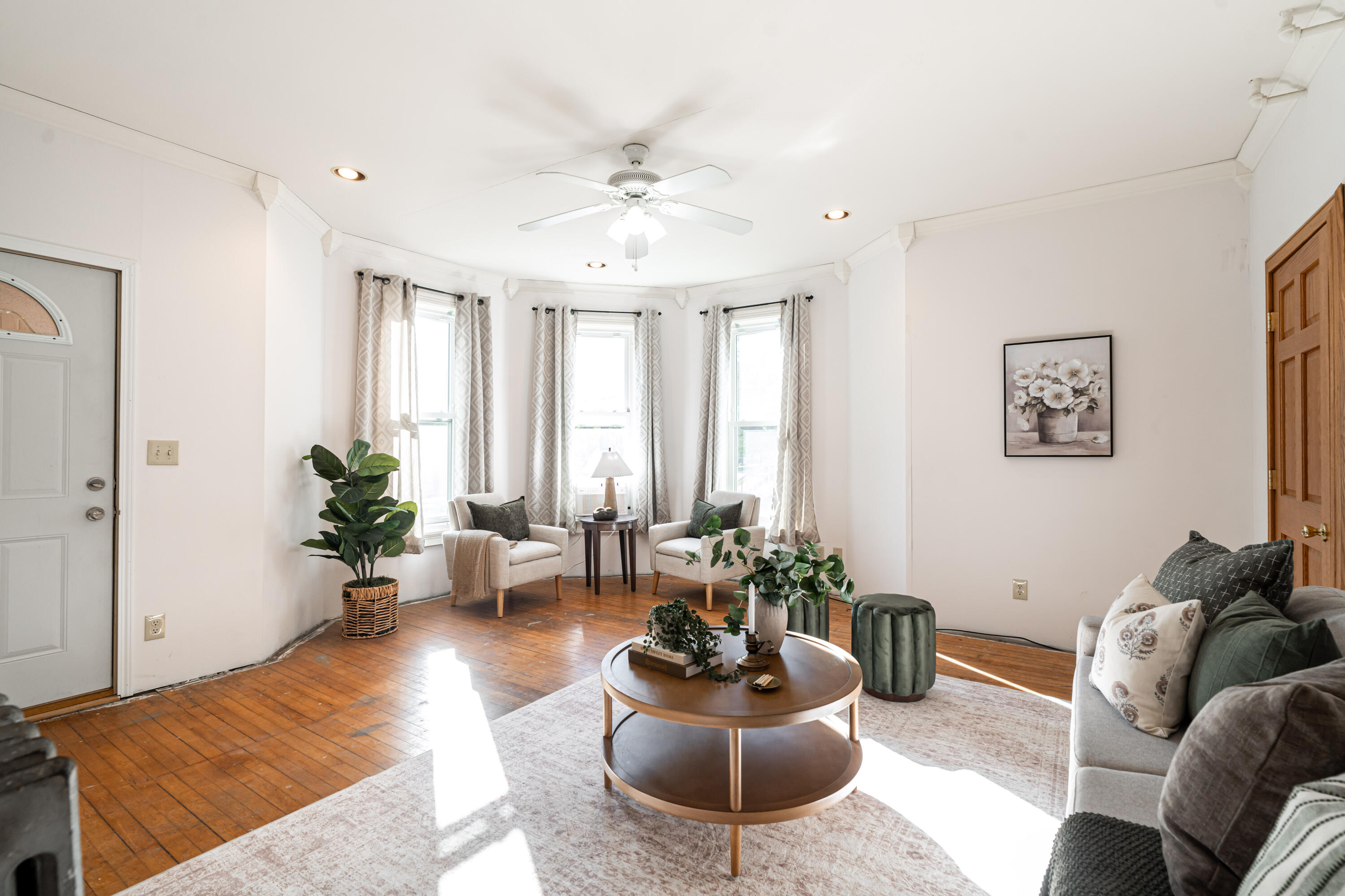 Living room with curtained windows, hardwood floors, and neutral seating around a round coffee table.