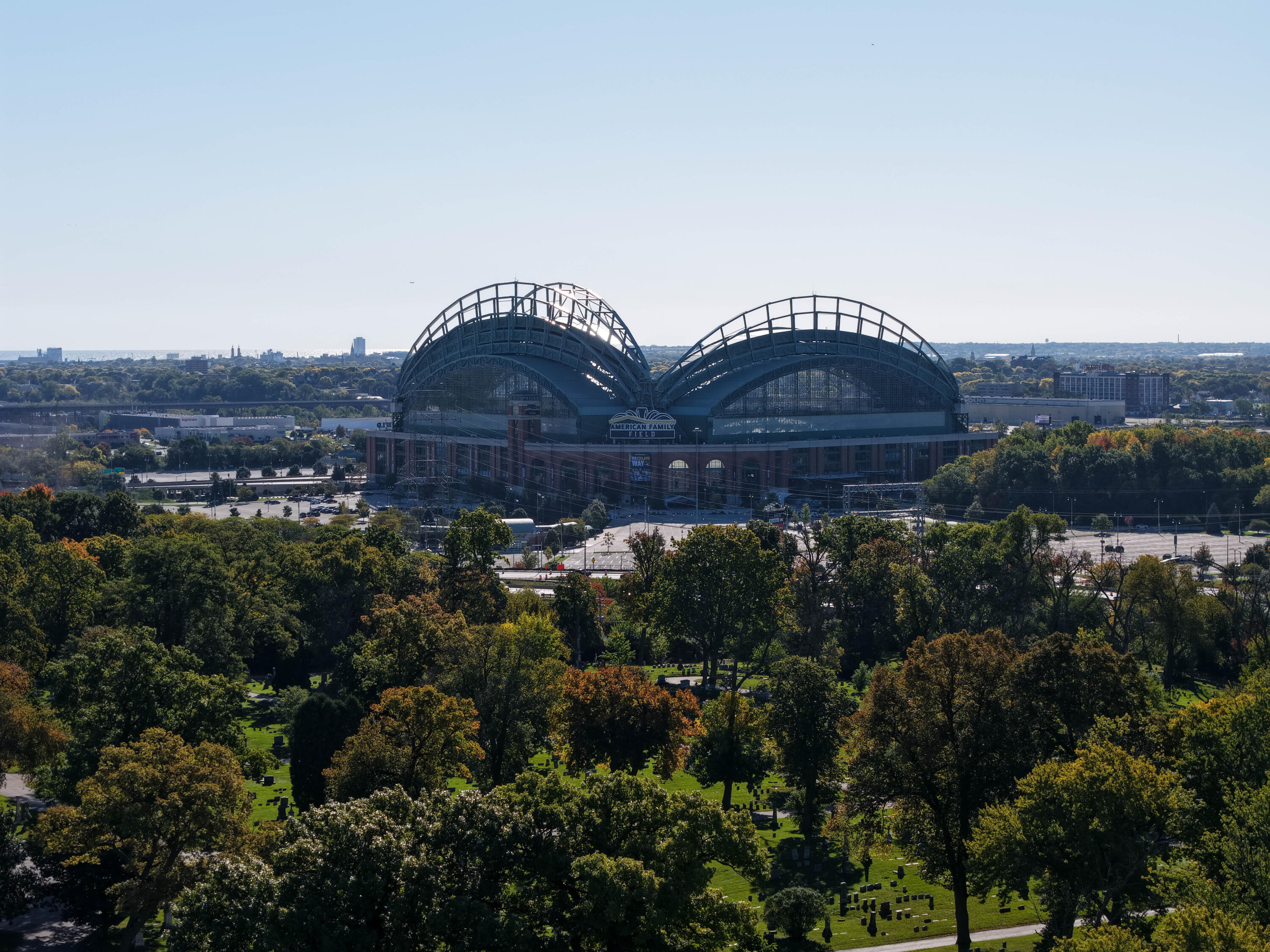 American Family Field rises above autumn trees, with a cemetery in the foreground and Milwaukee’s skyline beyond.