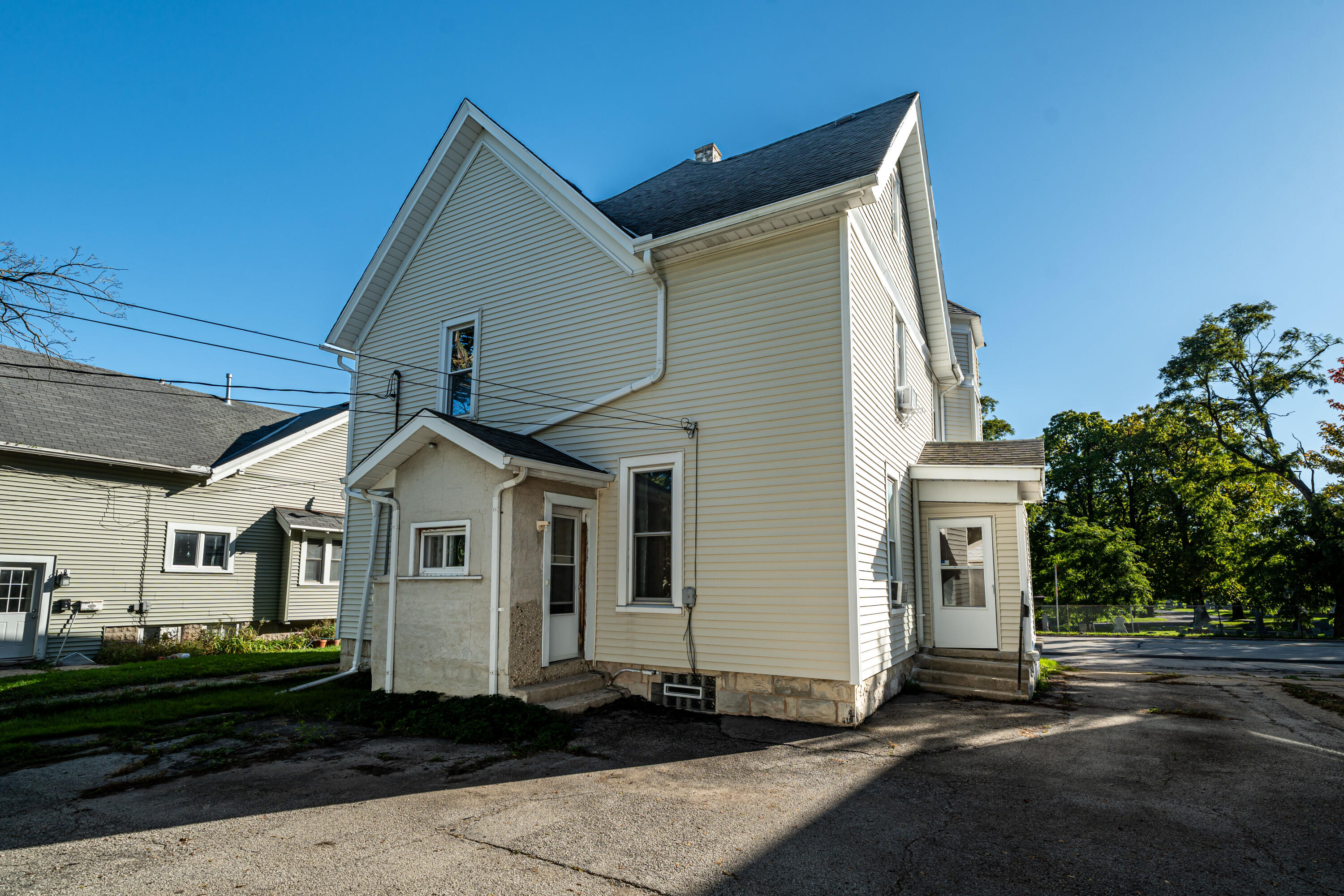 Side and rear of a two-story cream-sided house with a steep gabled roof and paved driveway.
