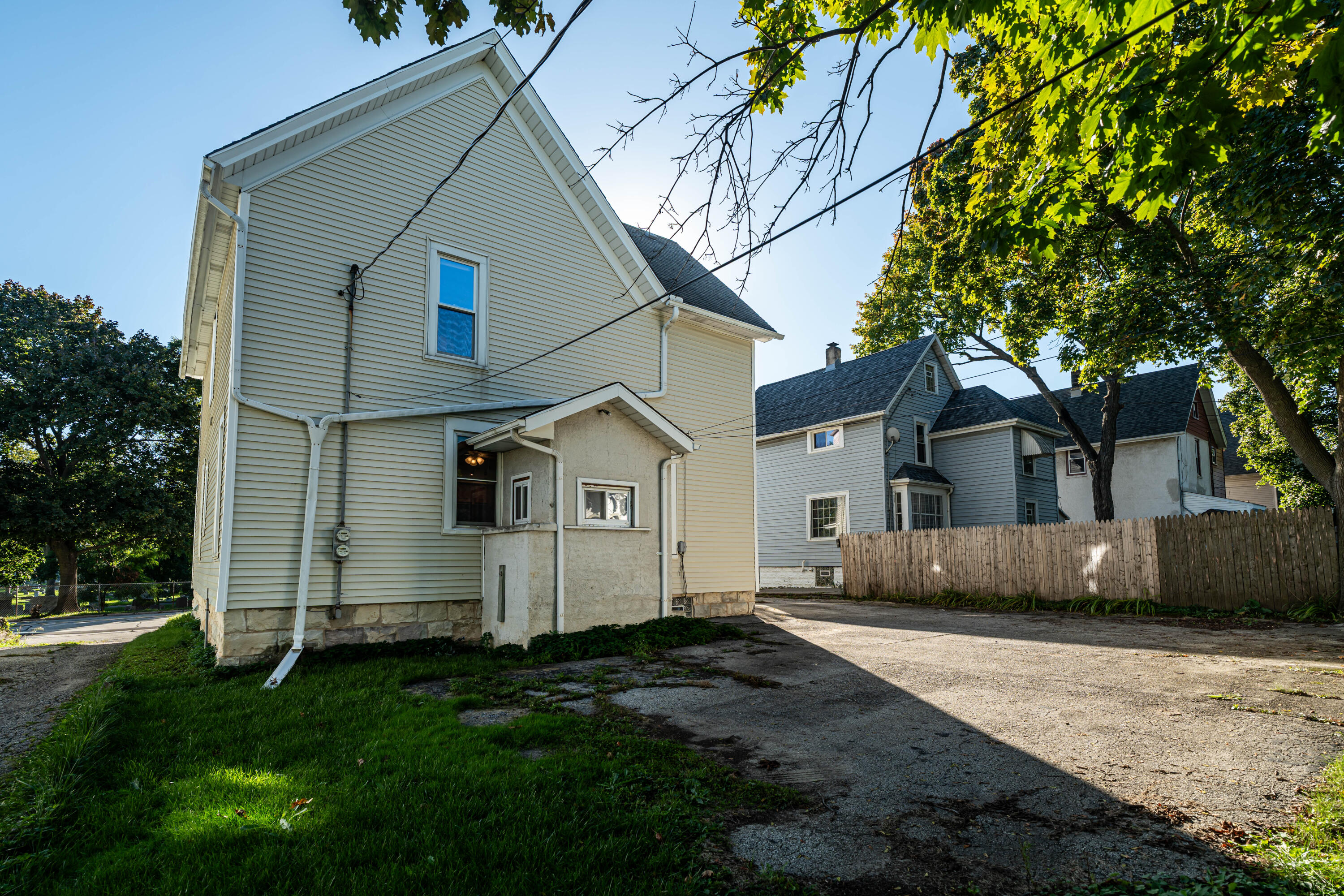 Two-story home with pale siding and a small enclosed rear entry beside a paved driveway and wooden fence.