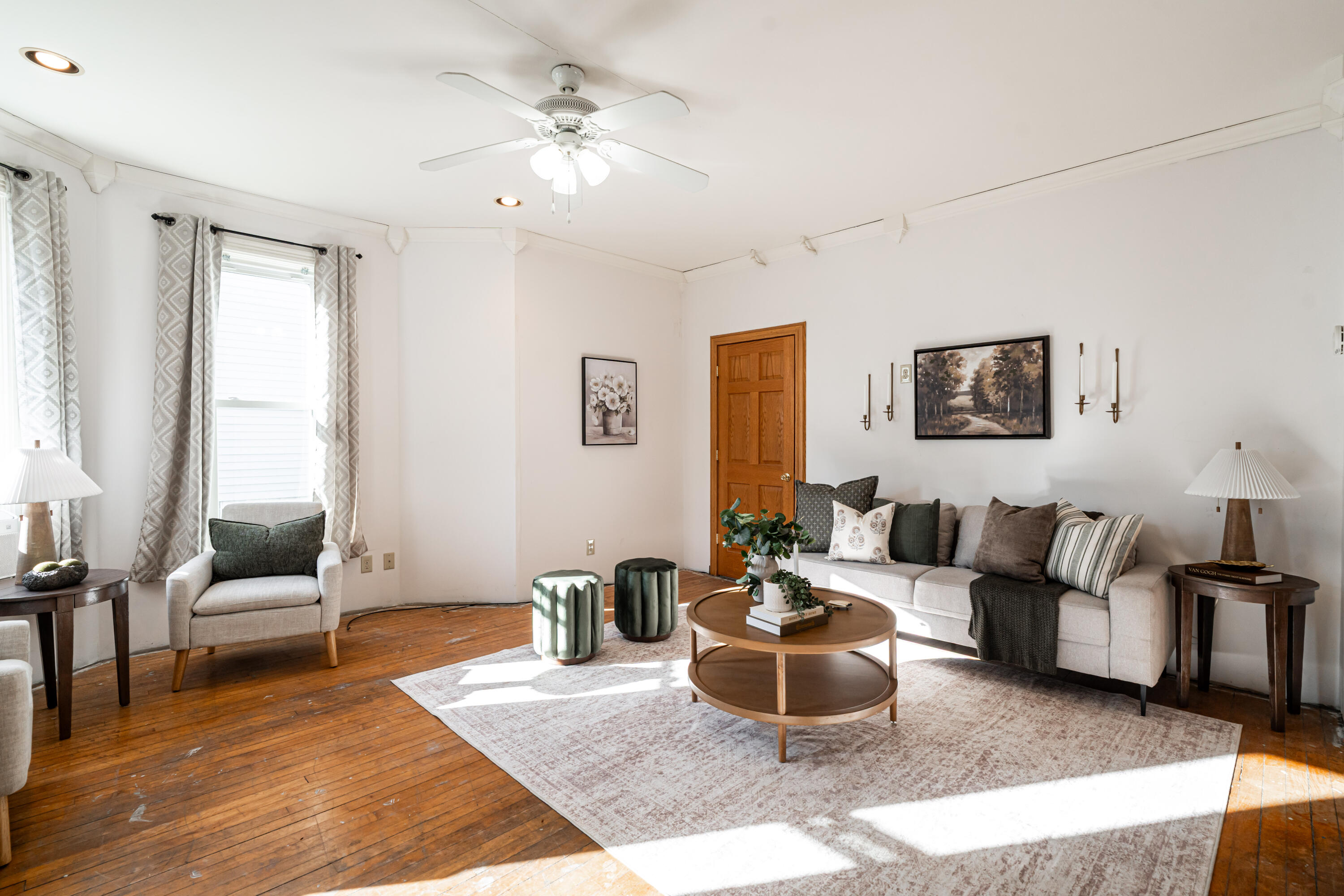 Staged living room with a light sofa, round coffee table, patterned rug, and sunlit hardwood floors.