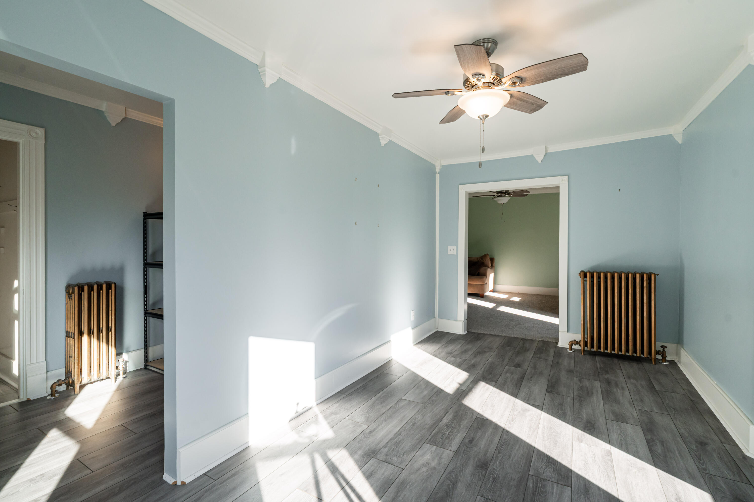 Empty blue room with gray plank flooring, ornate white trim, ceiling fan, and cast-iron radiators.