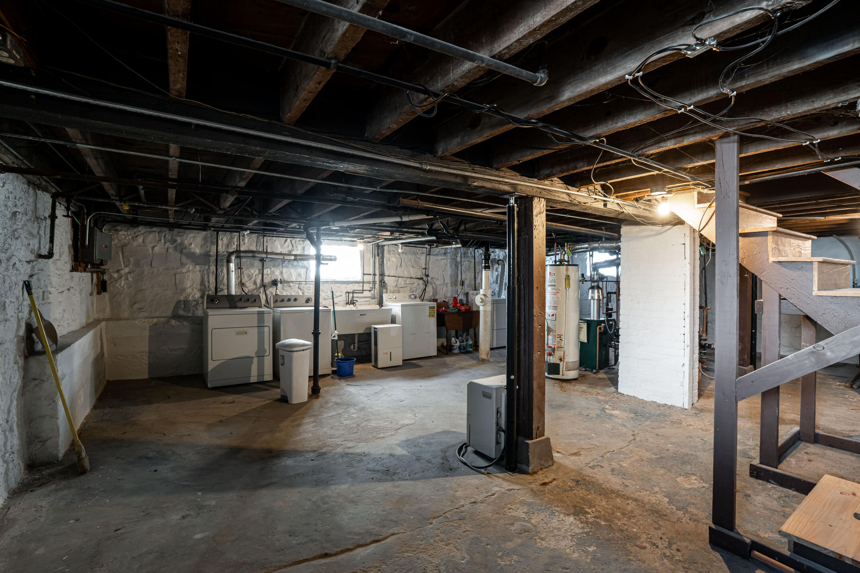 Basement laundry area with exposed beams, stone walls, washer and dryer, and a wooden staircase.