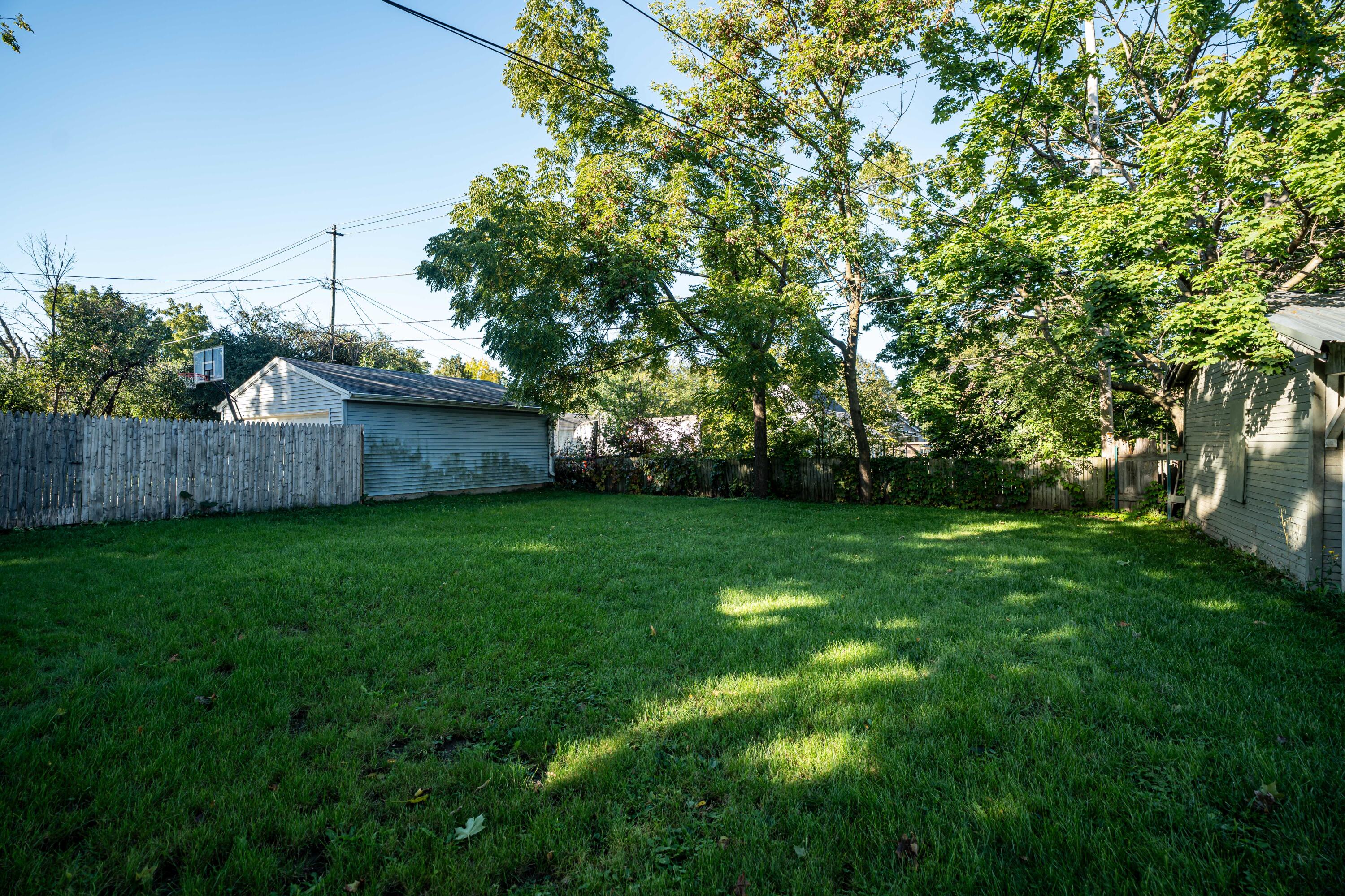 Grassy backyard with mature trees, fenced lawn, basketball hoop, and detached garage.