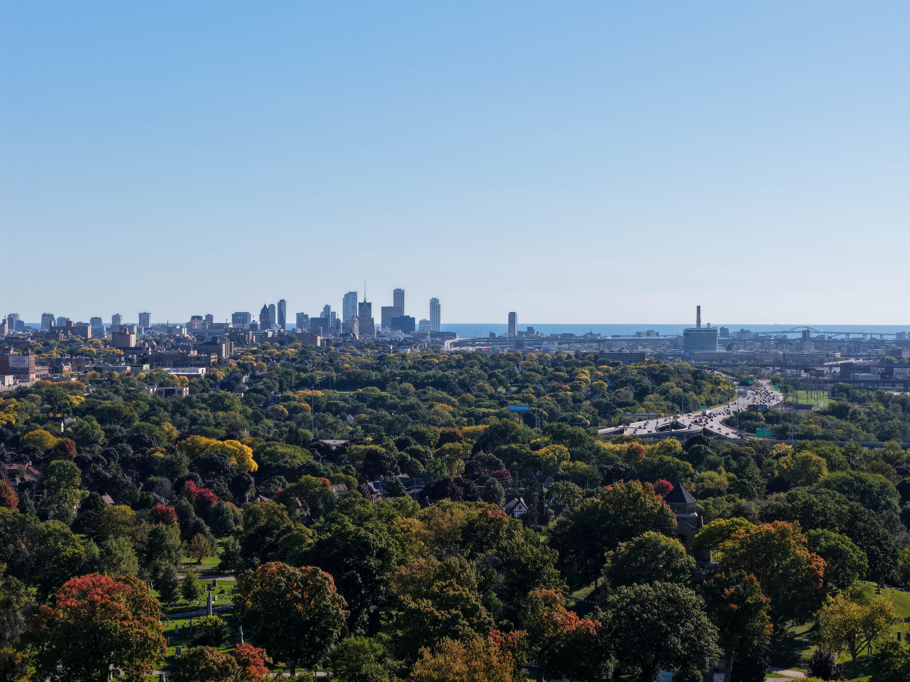 Milwaukee skyline and Lake Michigan beyond autumn-colored neighborhood trees and a curving highway.