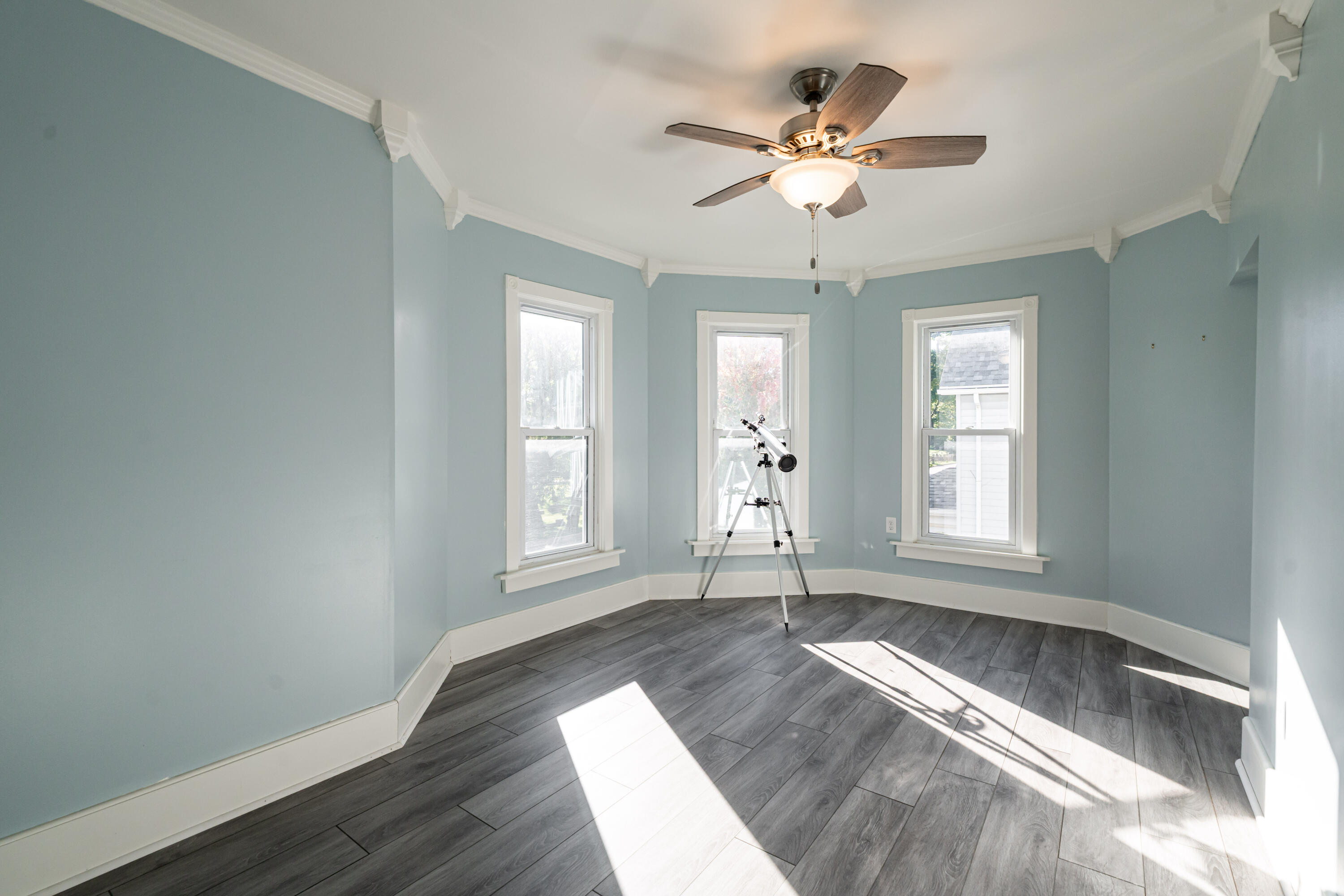 Vacant blue-walled room with a telescope between three windows and gray plank flooring.