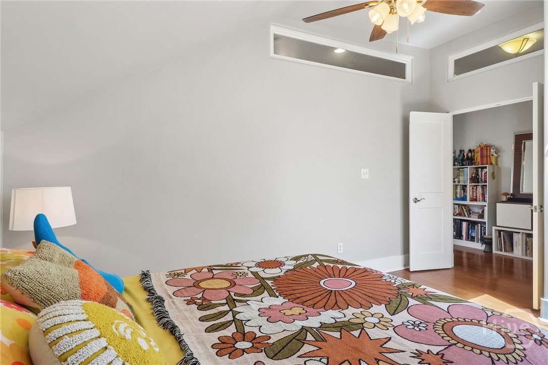 Bedroom with a colorful floral bedspread, ceiling fan, and open doorway to a book-filled room.
