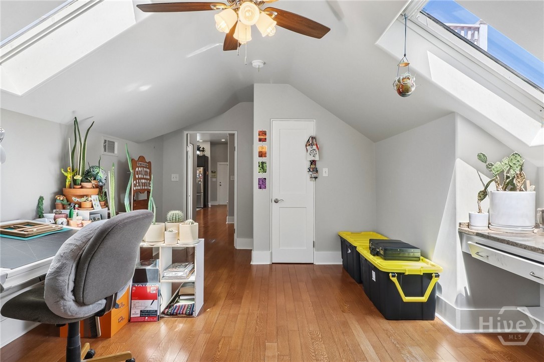 Attic office with vaulted ceiling, skylights, hardwood floors, desk, plants, and storage bins.
