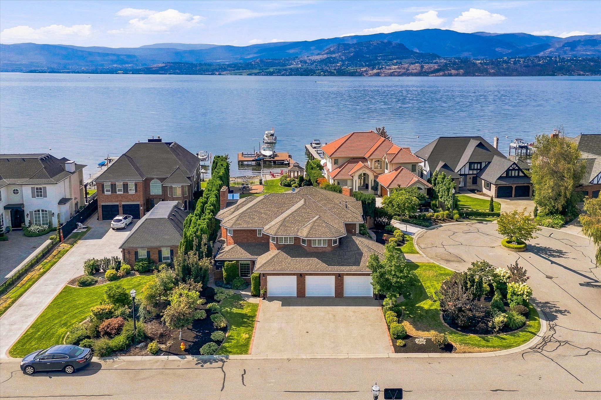 Aerial view of a brick home and landscaped waterfront neighborhood beside a lake with mountain views.
