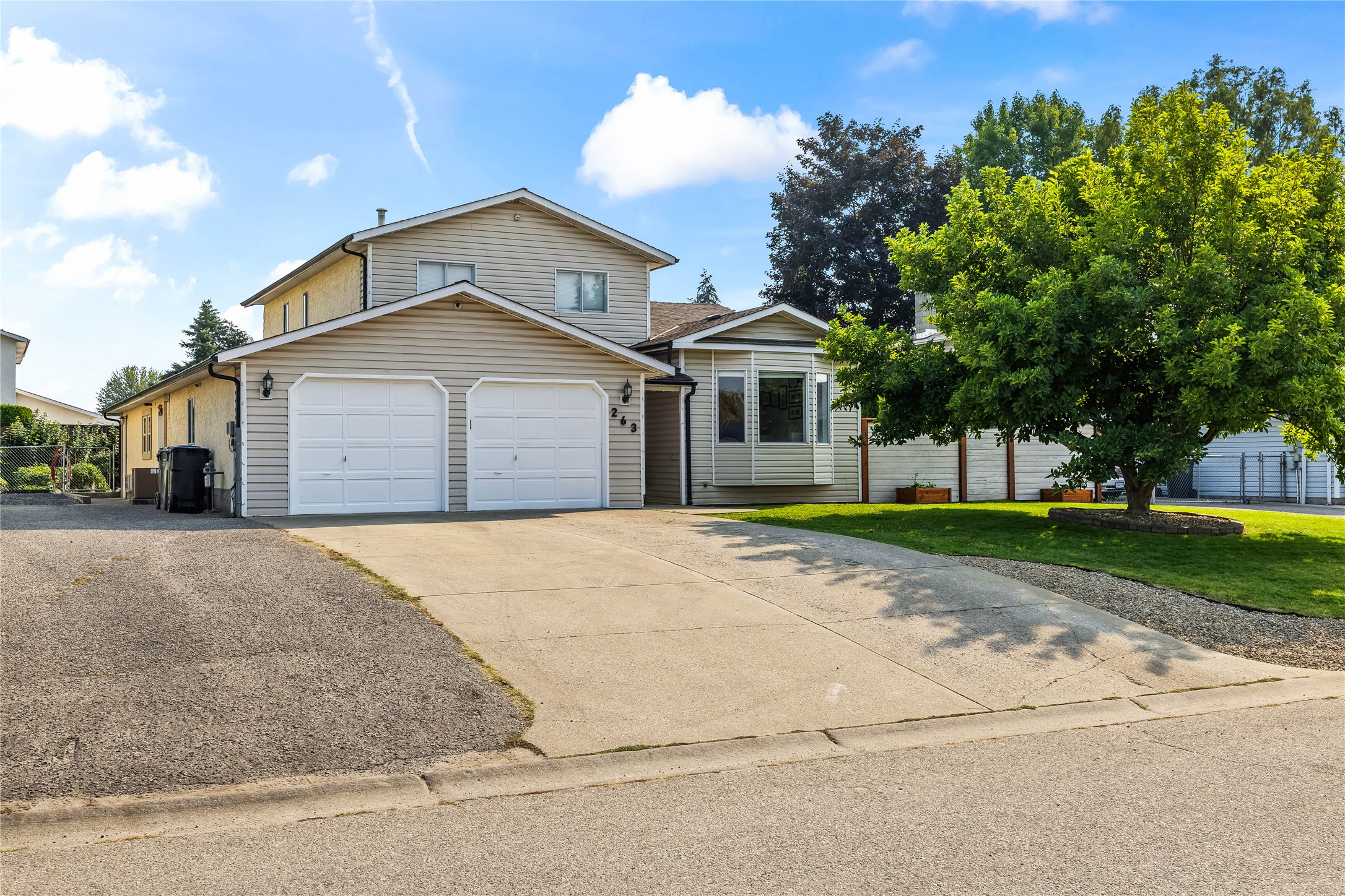 Two-story beige house with a double garage, wide driveway, and large leafy tree.