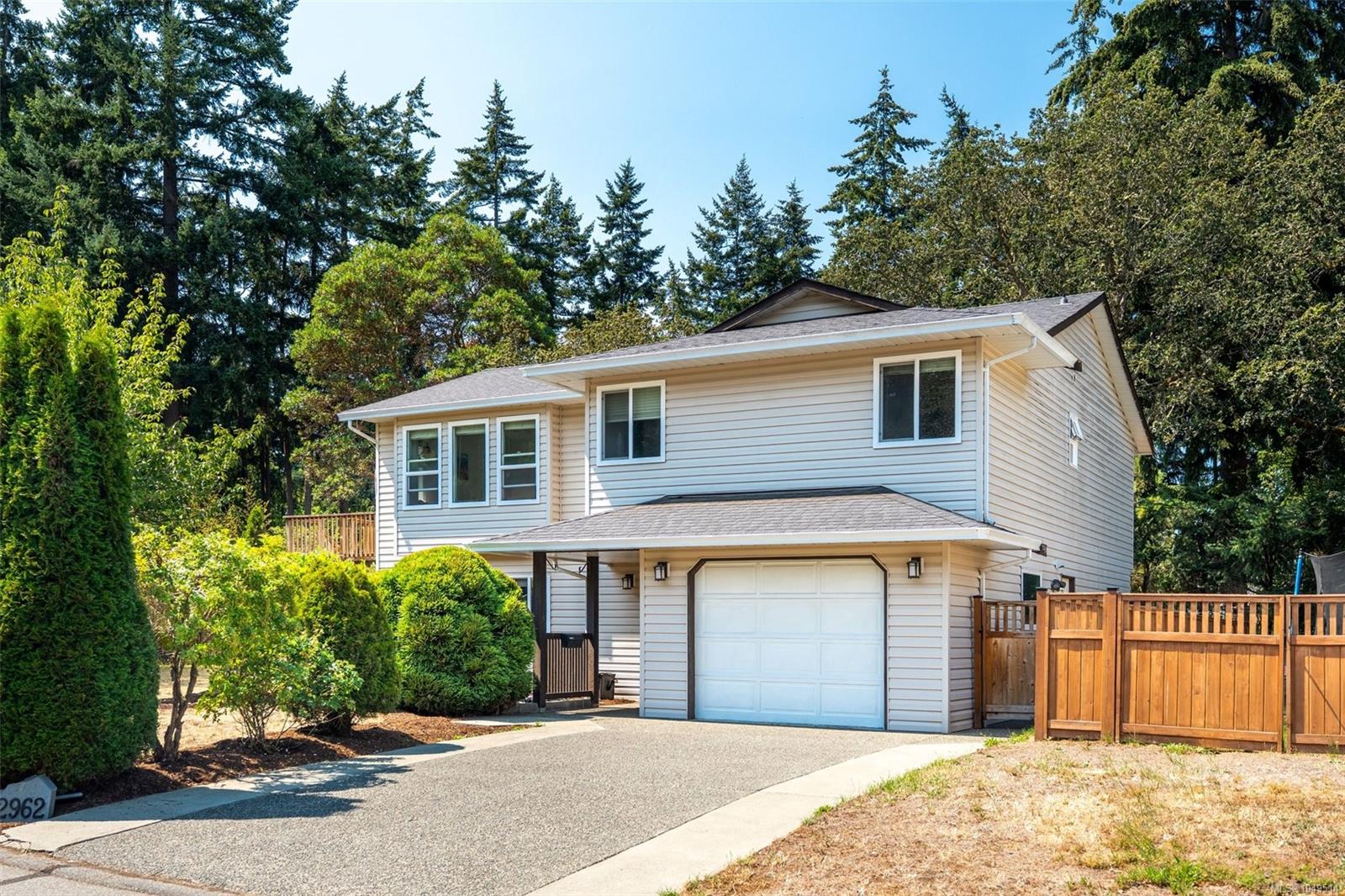 Two-story light-colored house with a white garage door, driveway, tall evergreens, and wooden fence.