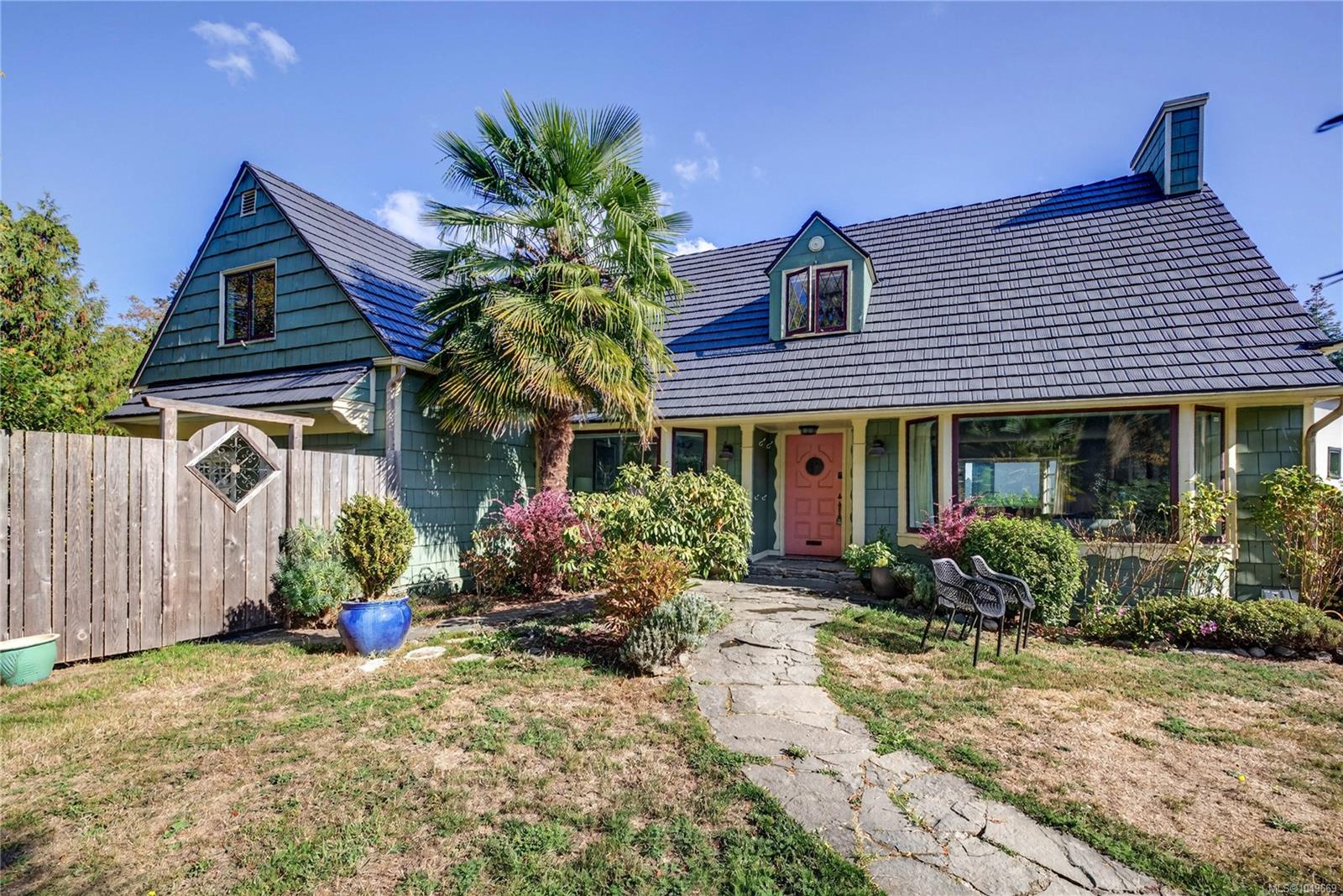 Teal shingle-sided house with a steep tiled roof, coral door, palm tree, and stone walkway.