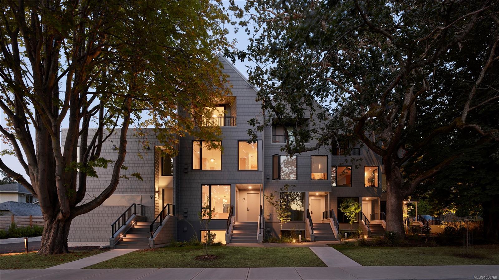 Modern townhouse exterior at dusk, framed by mature trees with warmly lit windows.